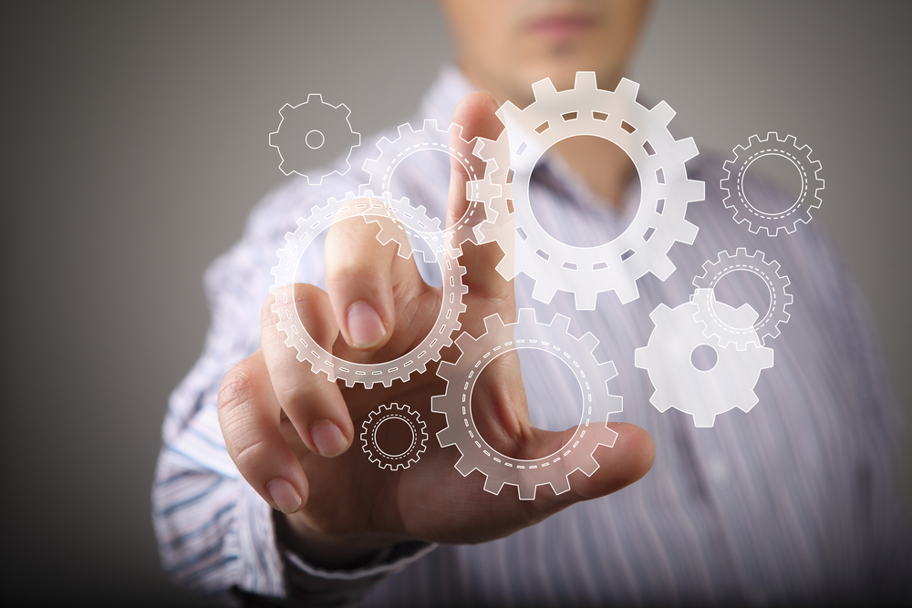 A man's hand is touching a gear wheel on a grey background.