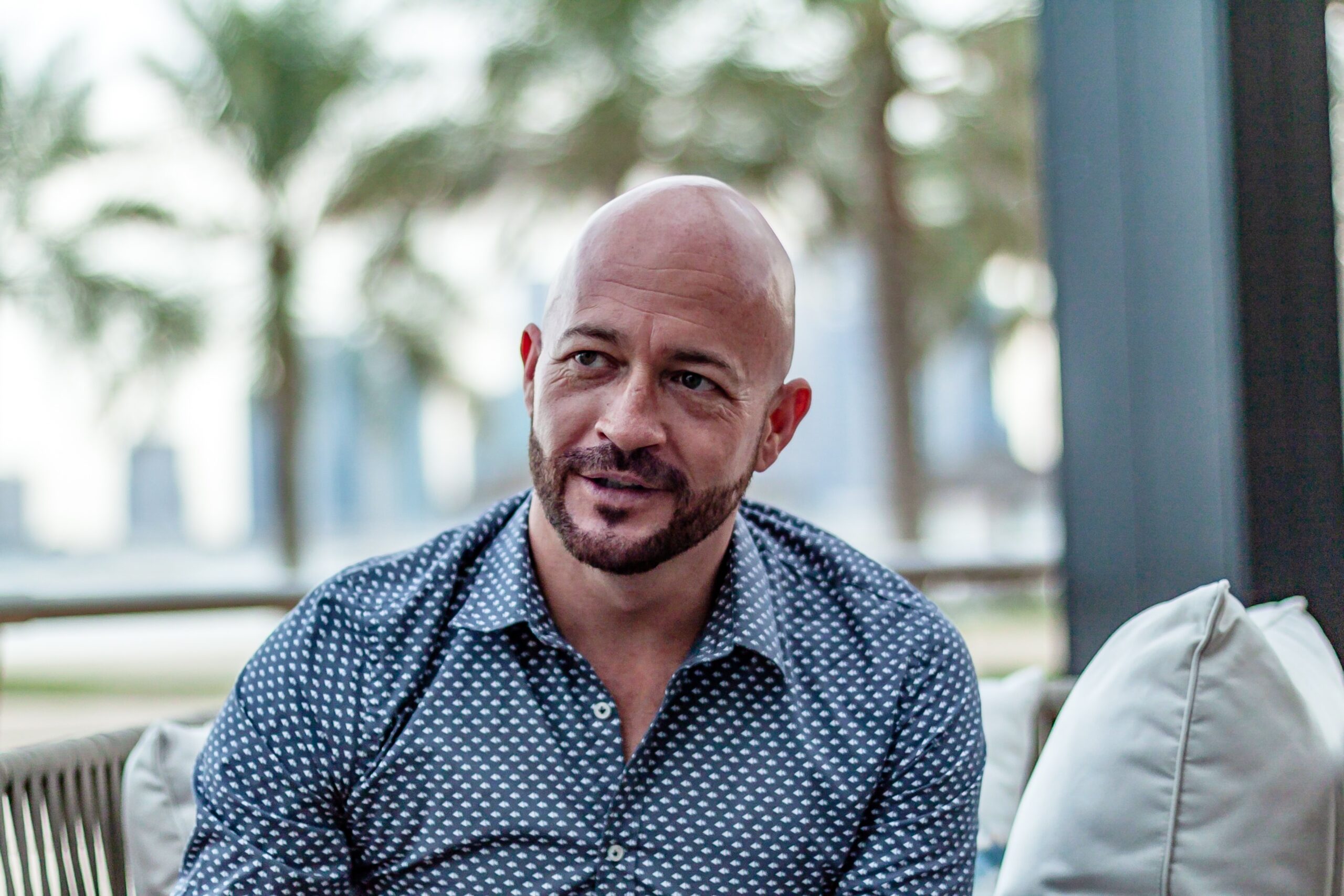 A bald man sitting on a couch with palm trees in the background.