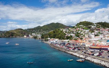 Aerial view of a coastal town with colorful buildings on a hillside. Boats are on the blue water and a road runs along the waterfront. Hills and fluffy clouds are in the background.