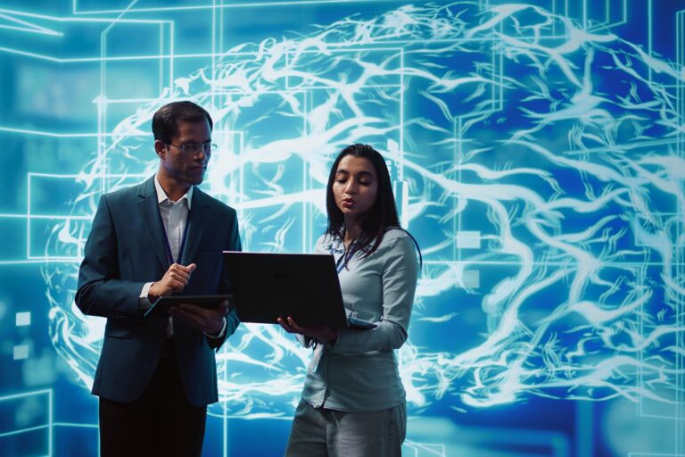 Two people stand discussing work with a laptop and tablet in hand, in front of a digital screen displaying a neural network or brain-like illustration.