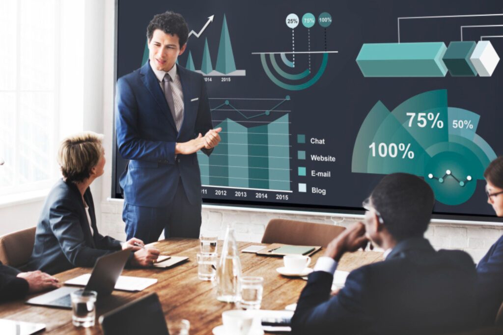 A man in a suit presents data charts on a screen, illustrating the benefits of CRM for small businesses to four colleagues seated at a conference table in a meeting room.