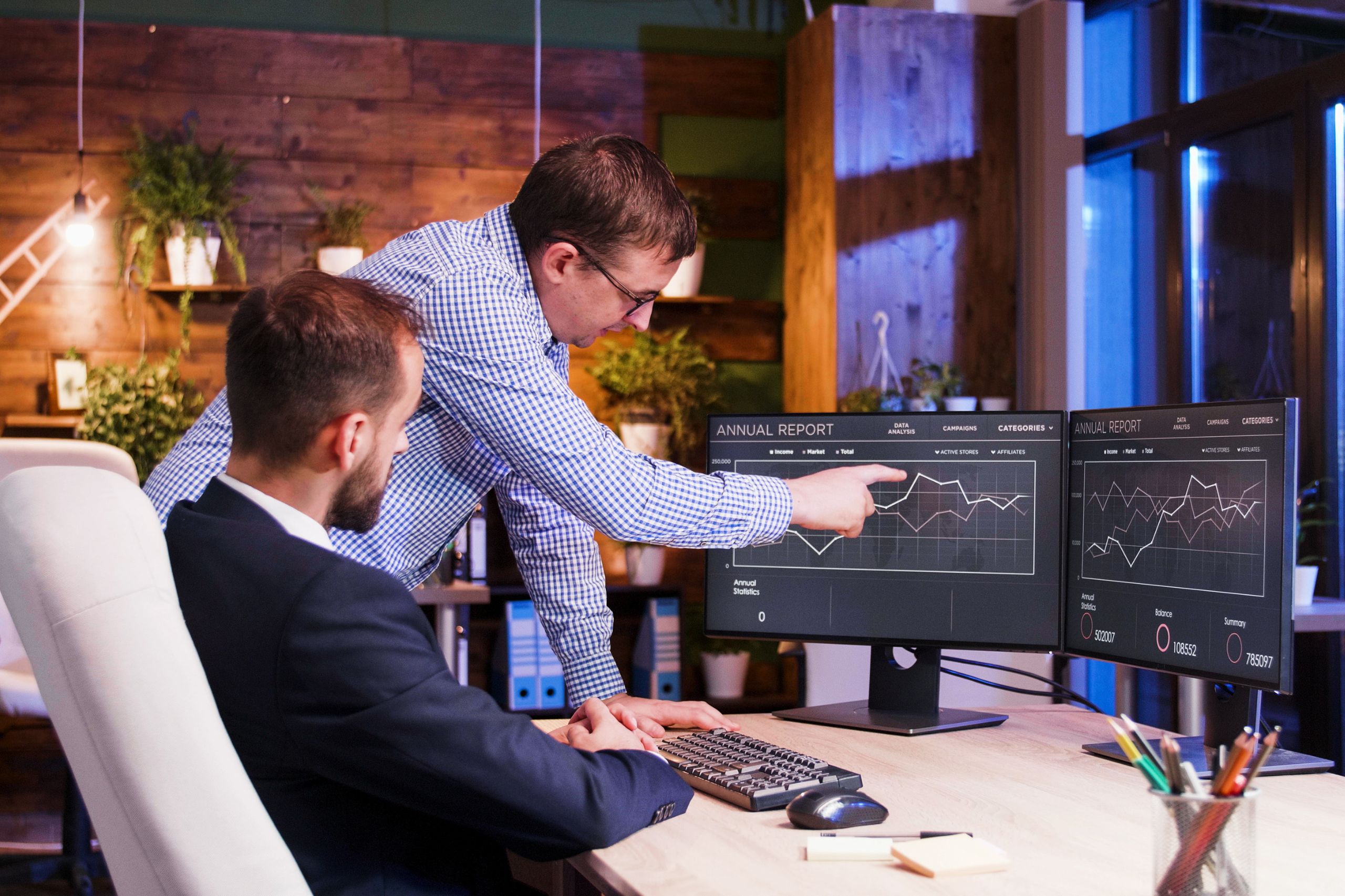 Two men in an office review graphs on computer monitors displaying an annual report, discussing IT leadership for SMEs as one points at the screen and the other listens.