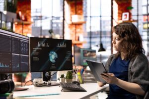 A woman sits at a desk in an office, holding a tablet and reviewing charts on two monitors, illustrating how marketing automation tools for SMEs streamline data analysis and improve business efficiency.