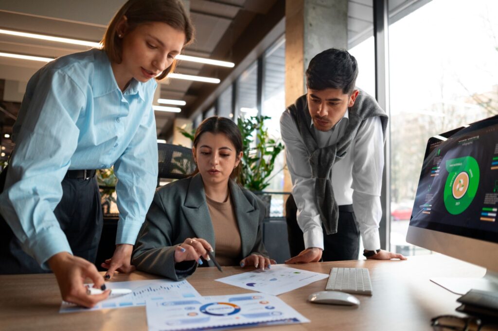 Three people in business attire review printed charts and graphs at a desk in an office, with a computer displaying a data dashboard nearby—highlighting SME project management strategies in action.