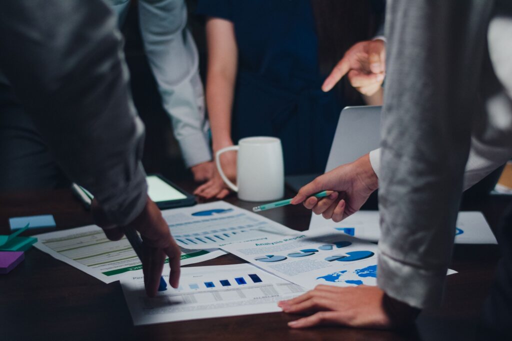 Several people stand around a table reviewing charts and graphs, discussing SME project management strategies, with hands pointing and gesturing among coffee mugs and electronic devices.
