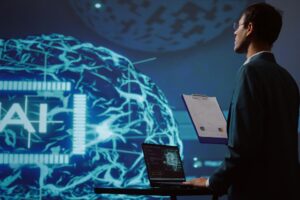 A man in a suit stands with a clipboard and laptop, facing a large digital display showing a neural network and the letters "AI.