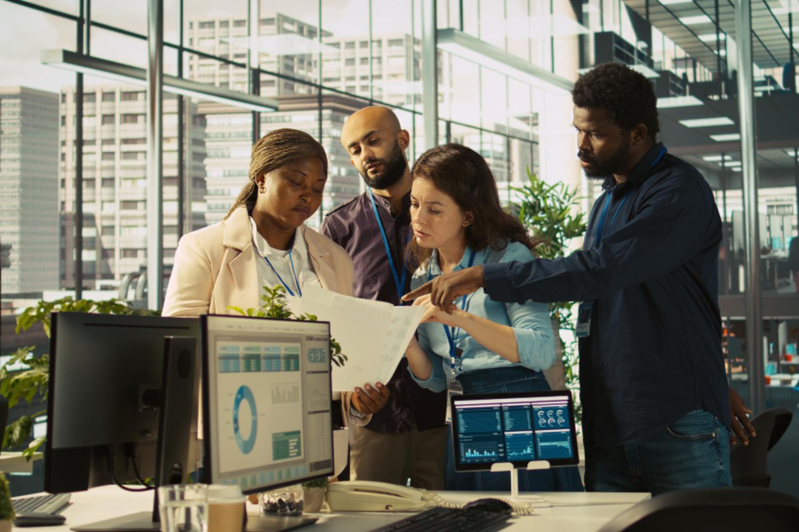 Four colleagues stand together in a modern office, reviewing documents and discussing data displayed on computer monitors.