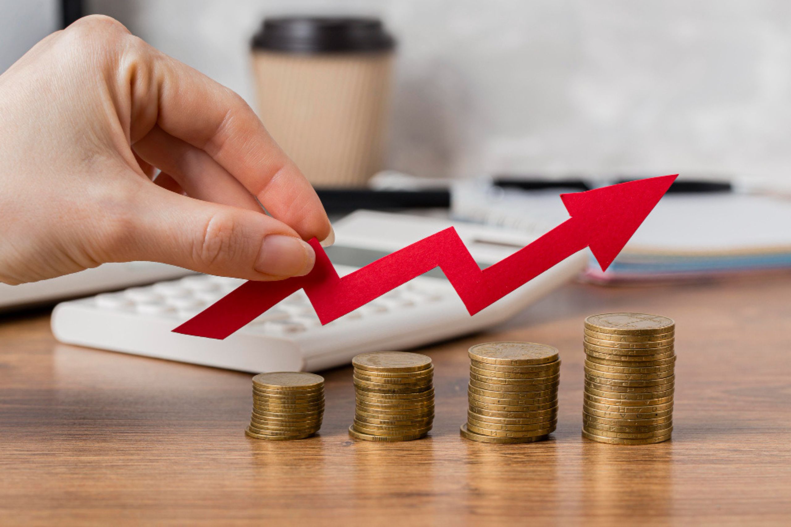 A hand holds a red upward arrow above four stacks of coins, symbolizing financial growth, with a calculator and coffee cup in the background.