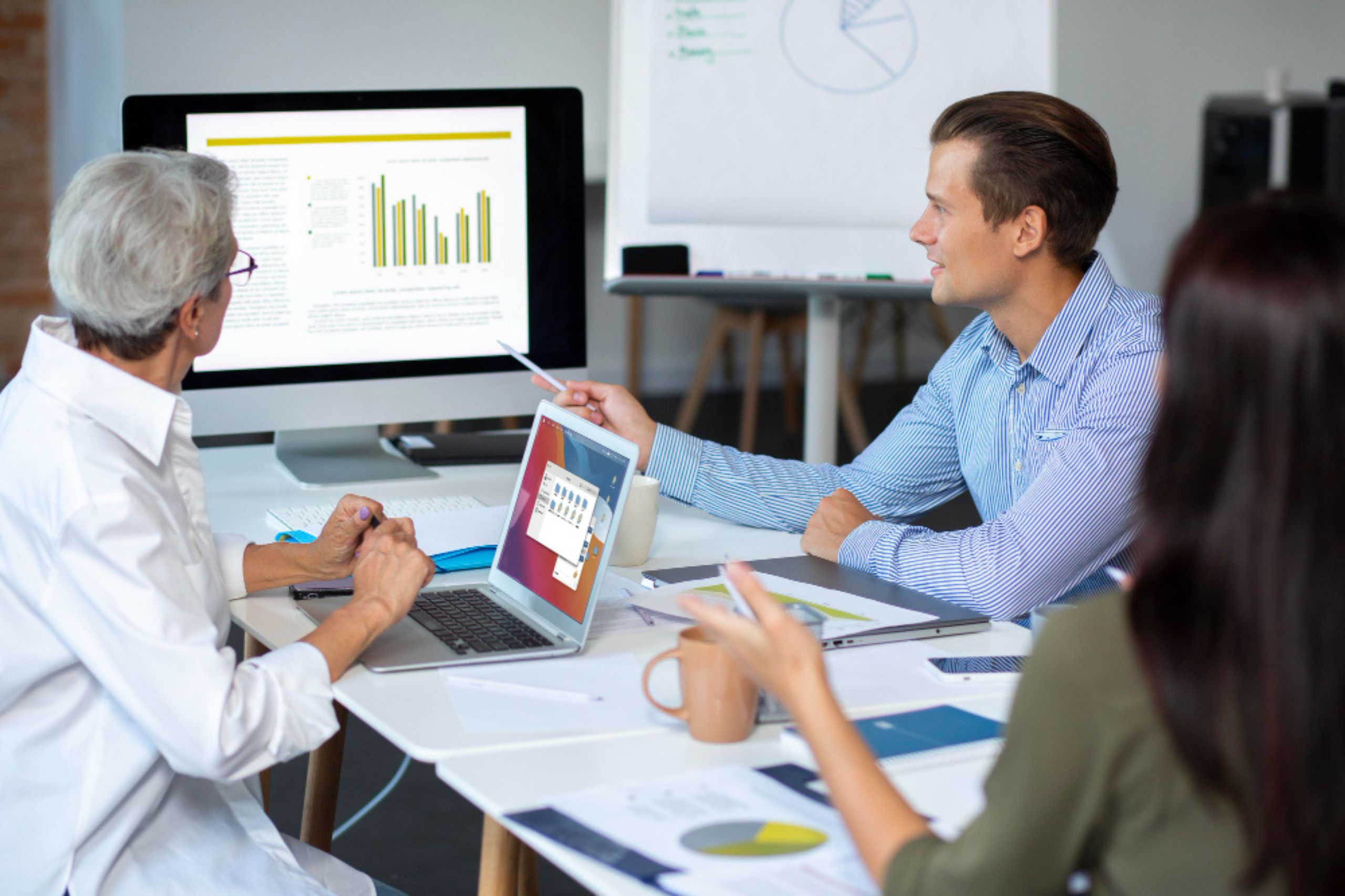 Three people sit at a desk reviewing charts and graphs displayed on a computer monitor and discussing data during a meeting.