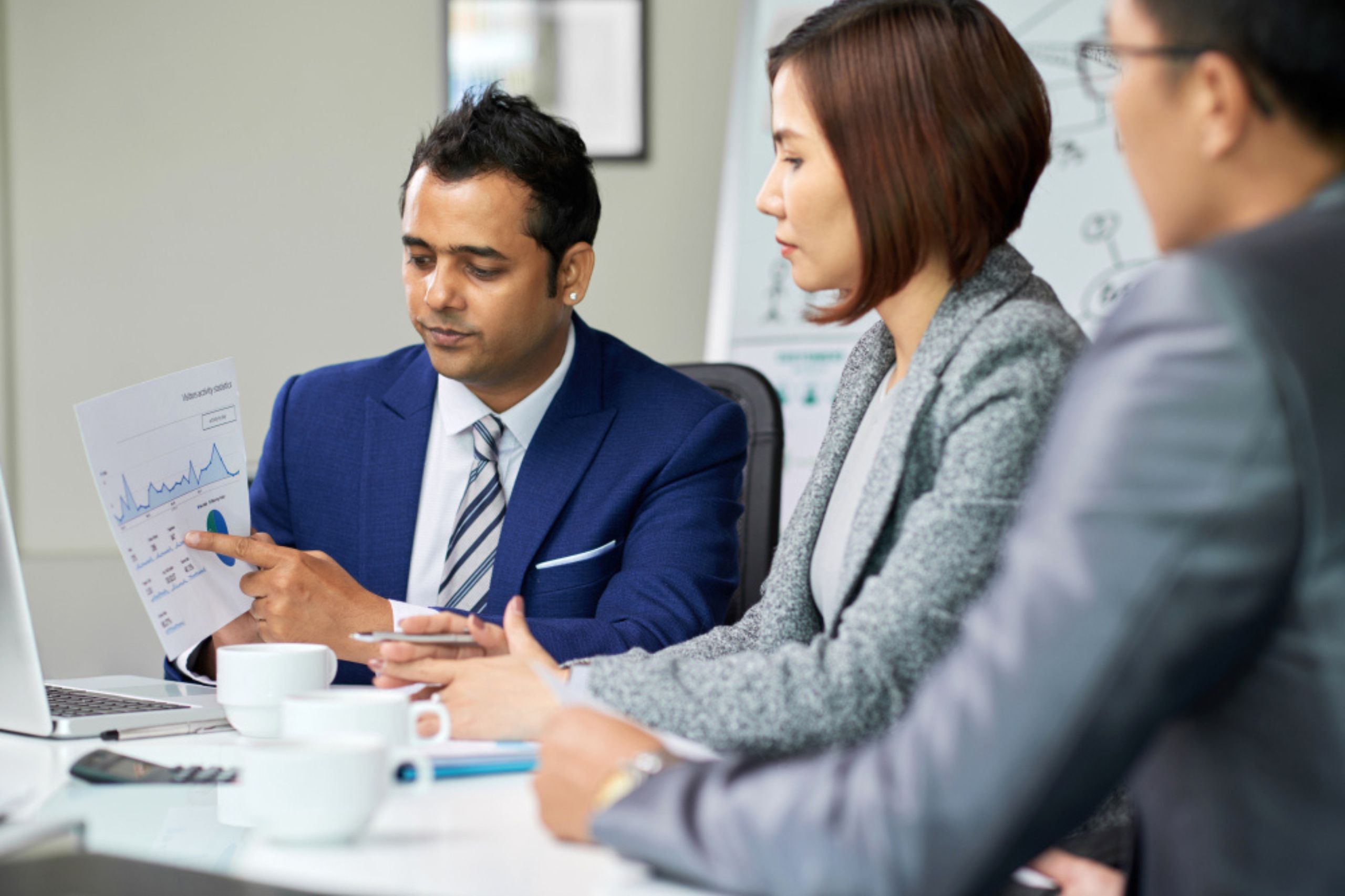 Three professionals in business attire discuss a printed graph at a meeting table with a laptop, documents, and coffee cups.