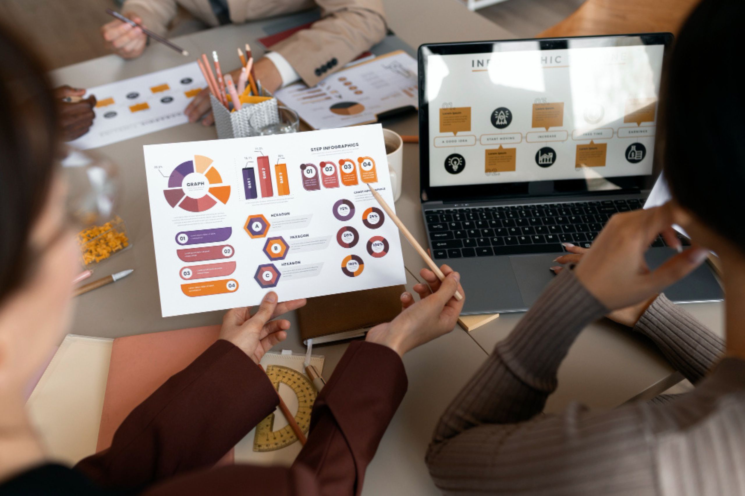 Two people sit at a desk reviewing a printed infographic and a laptop displaying a graph, surrounded by office supplies and charts, during a business meeting.