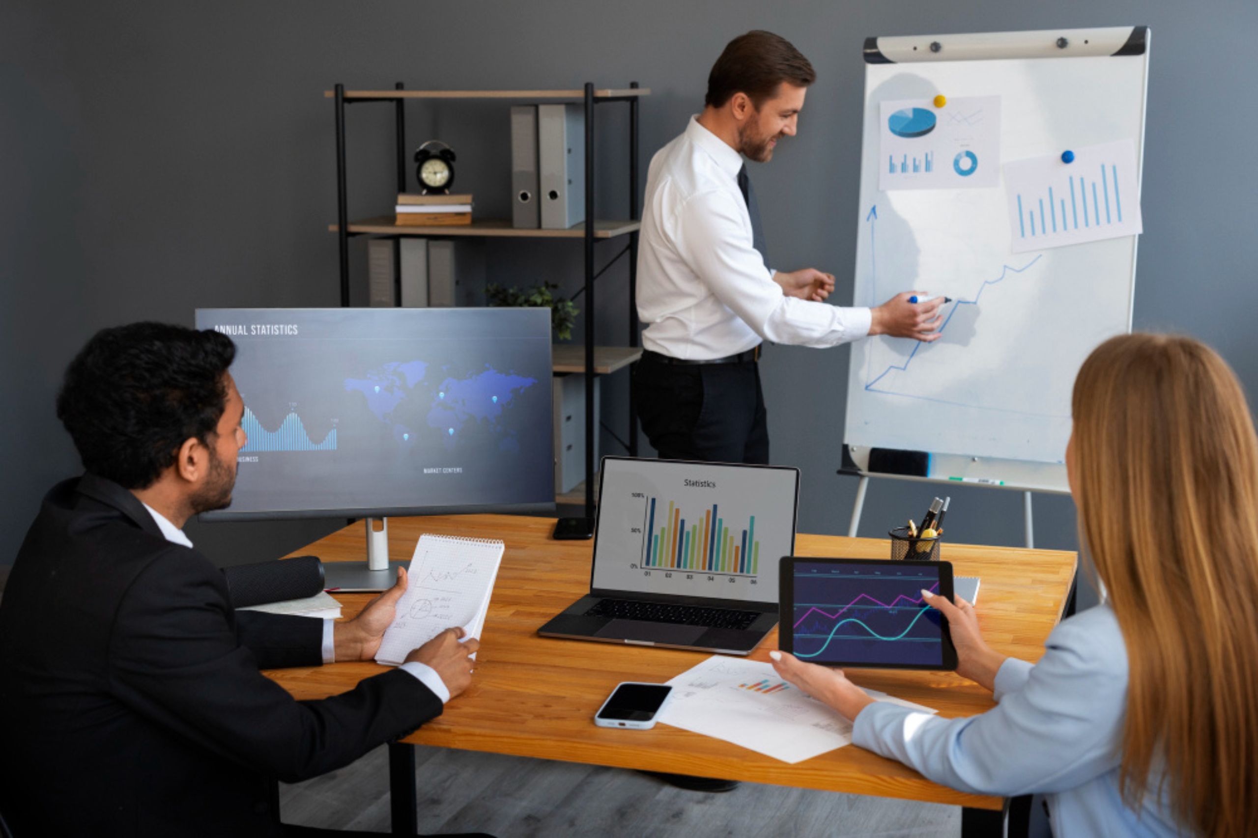 Three people in business attire review data and charts during a presentation; one person points at a line graph on a flipchart while others observe and take notes.