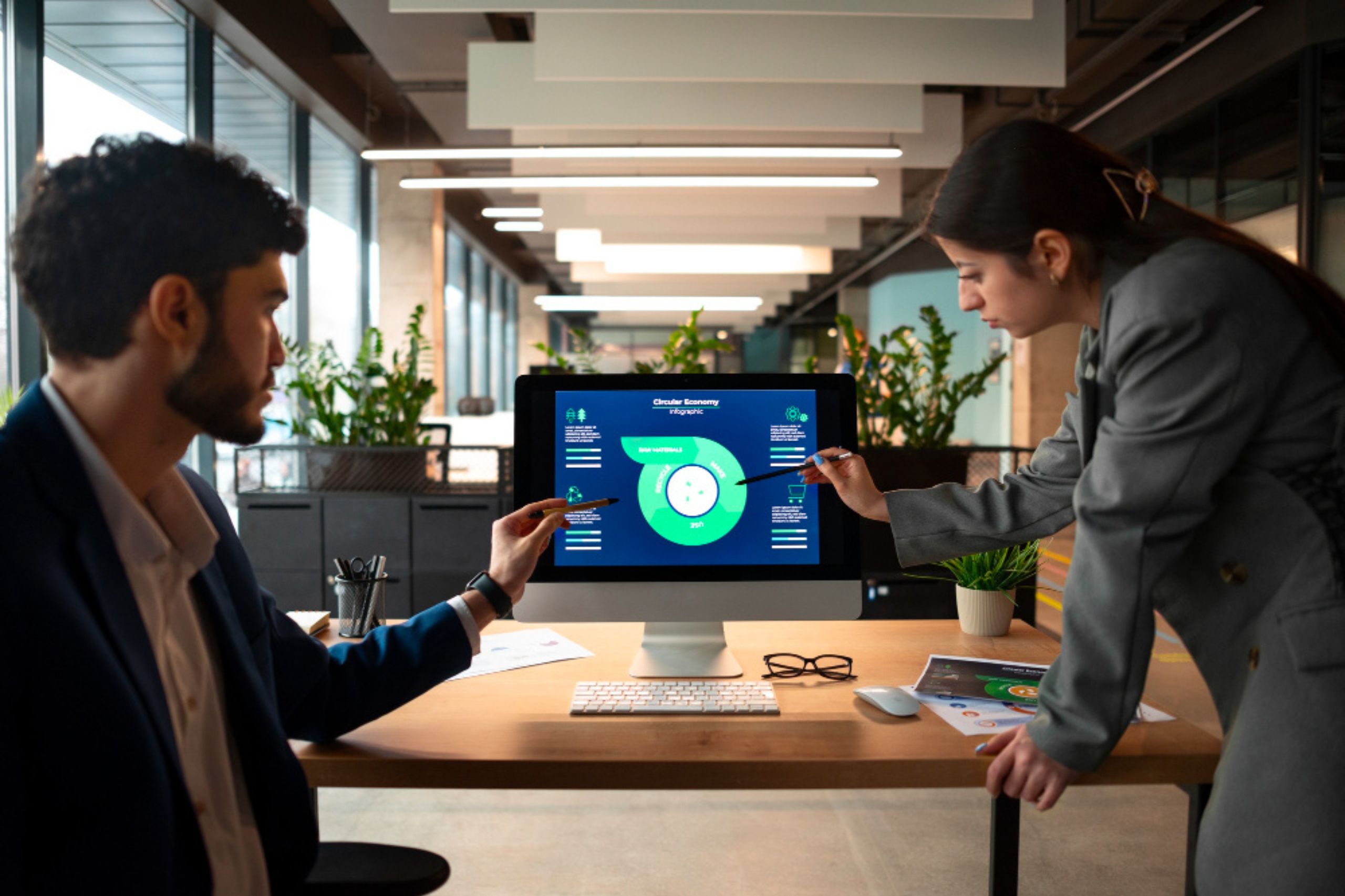 Two people in business attire discuss data displayed on a computer monitor in a modern office setting, pointing at a circular infographic on the screen.