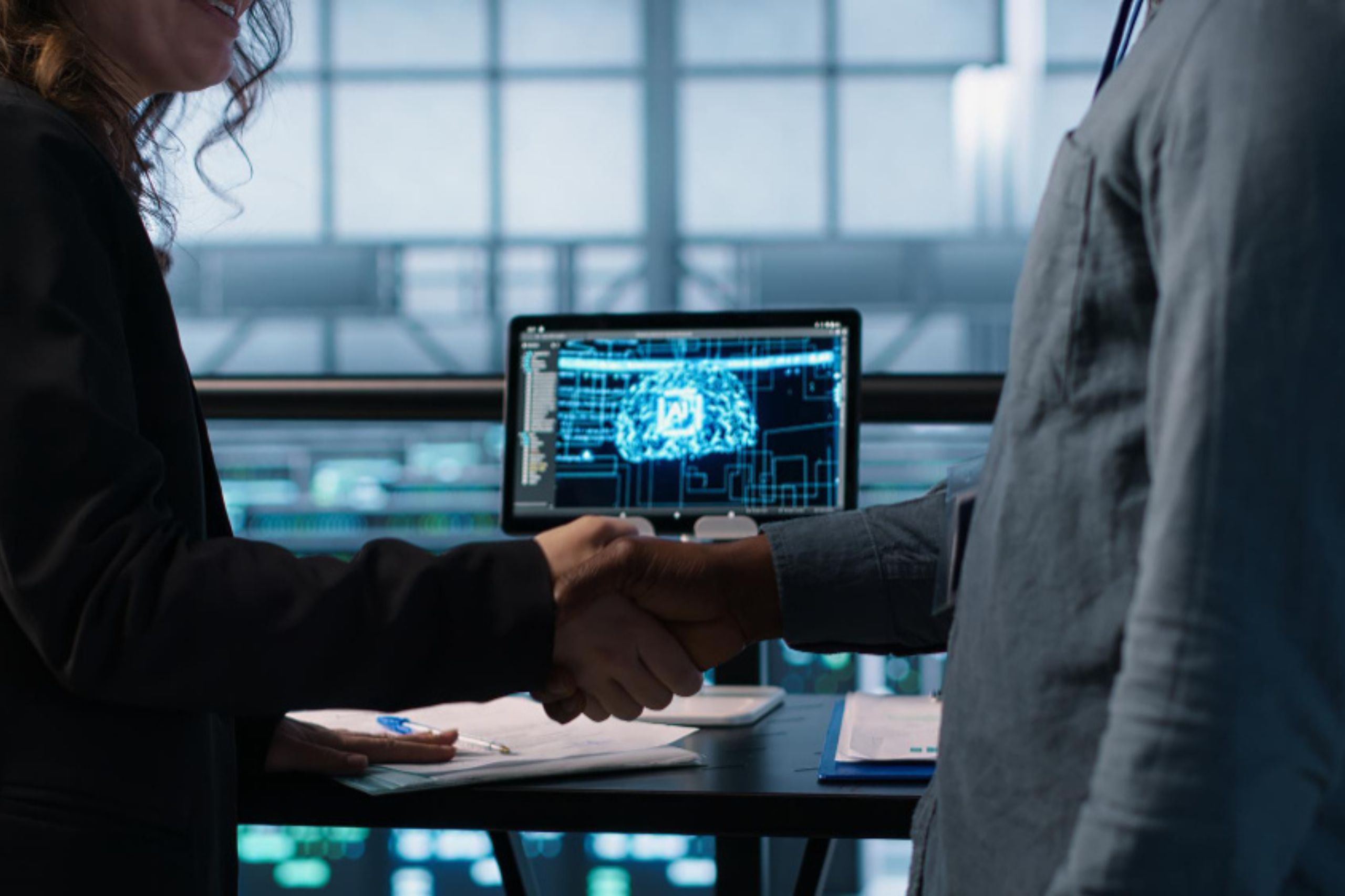 Two people in business attire shake hands in front of a computer displaying a digital network diagram, with documents and a pen on the desk.