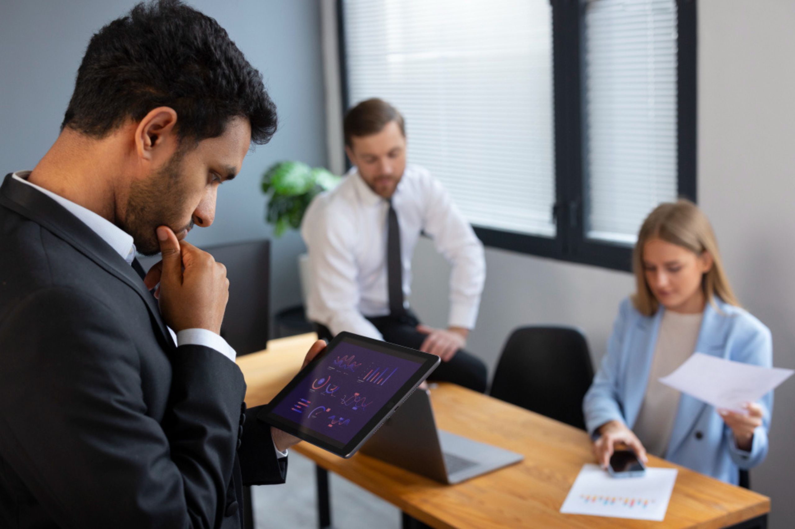 A man in a suit looks at data on a tablet while two colleagues sit at a desk with a laptop and papers in a modern office.