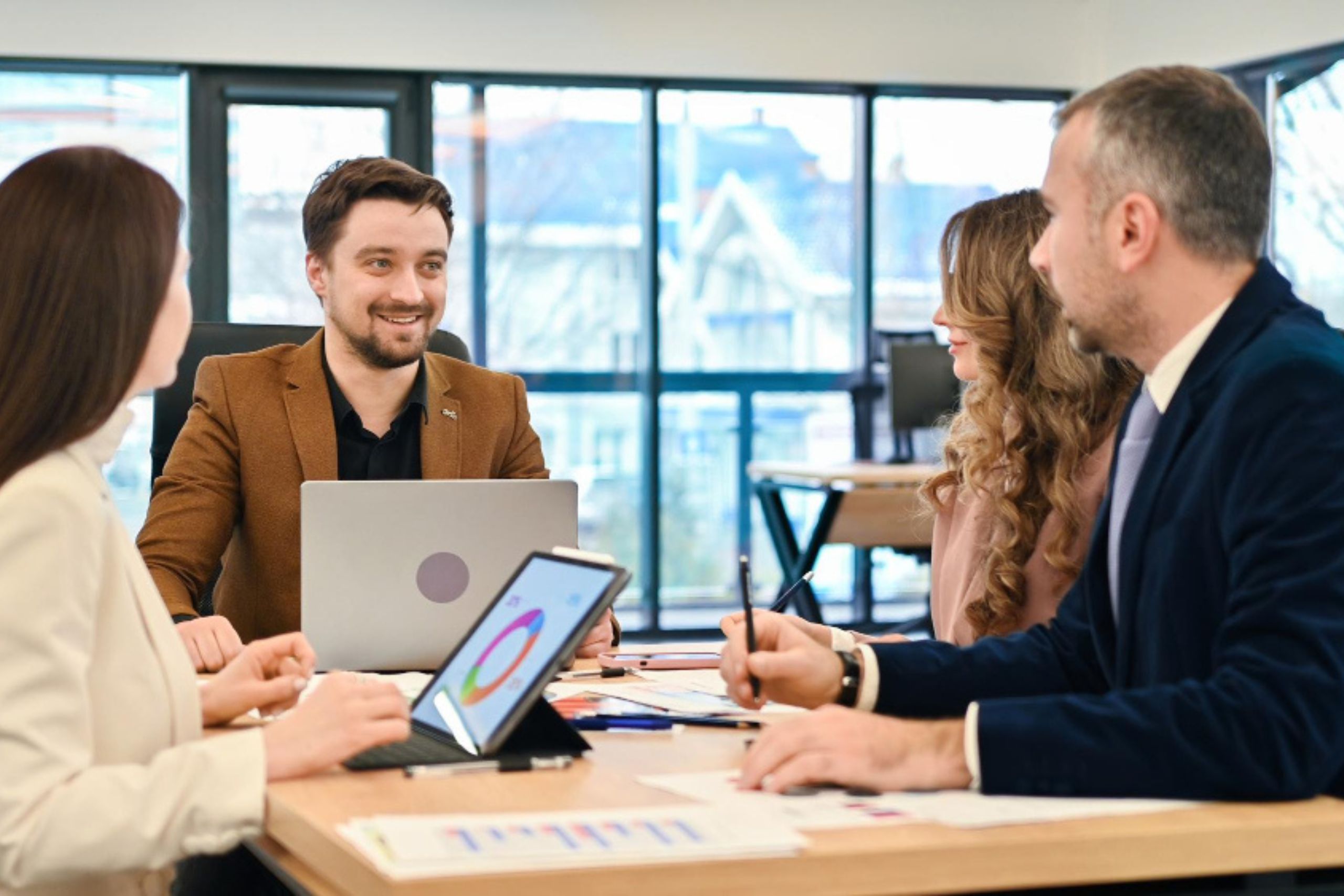 Four people sit around a table in an office, discussing documents and charts, with laptops and tablets in front of them. Large windows are visible in the background.