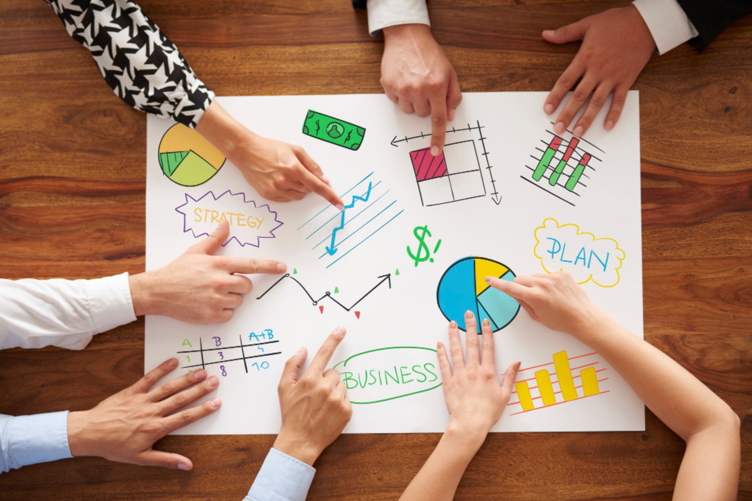 Six hands point to different graphs, charts, and business strategy illustrations on a large white paper, representing teamwork in planning and analysis on a wooden table.