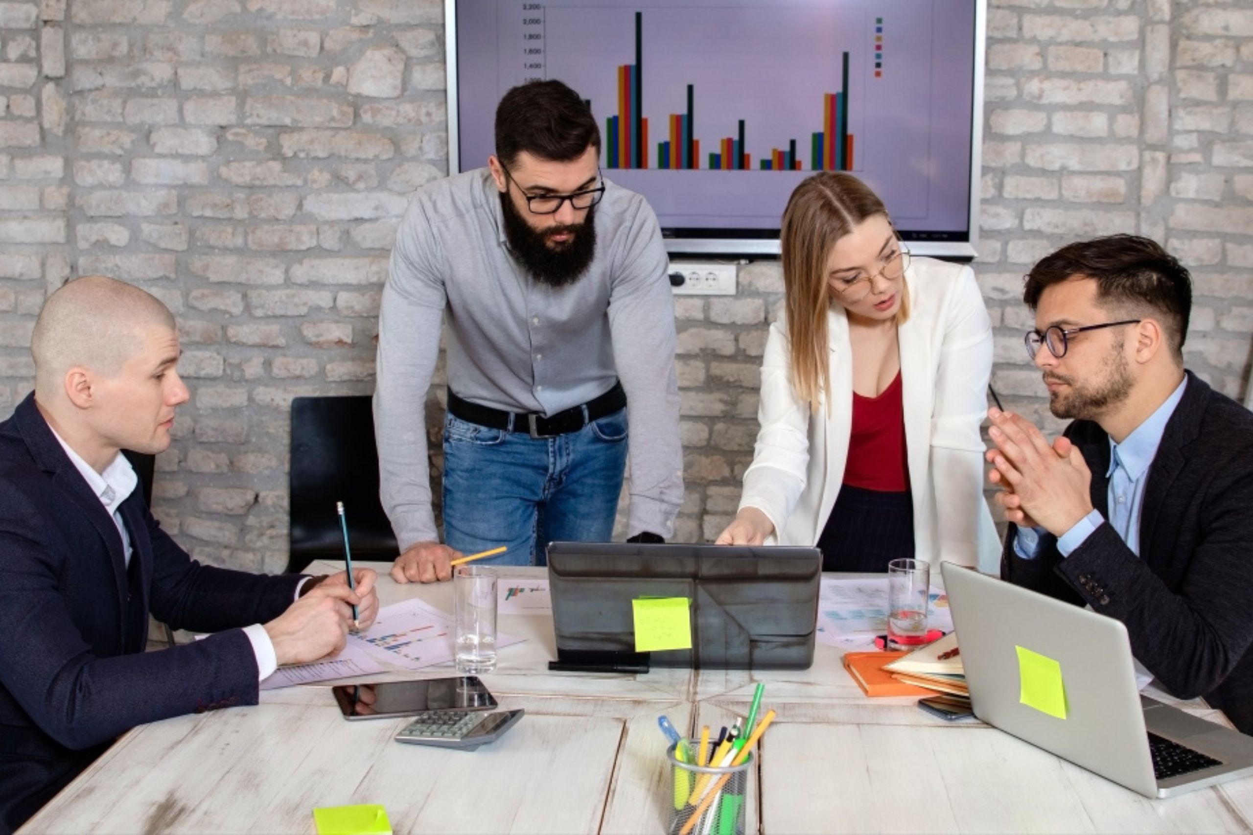 Four people in business attire have a discussion at a meeting table with laptops and documents, while a bar graph is displayed on a screen in the background.