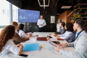 A man stands giving a presentation to six seated colleagues in a modern conference room, with a world map and data chart displayed on a screen.