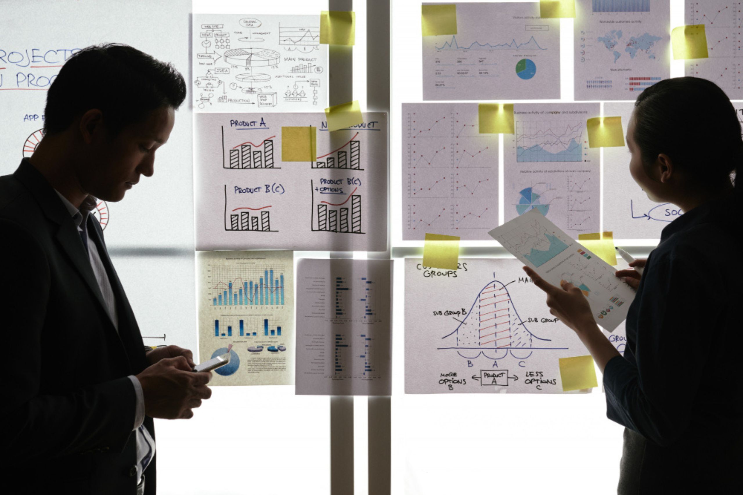 Two people look at documents and charts posted on a glass wall, discussing data and project plans in a brightly lit office setting.