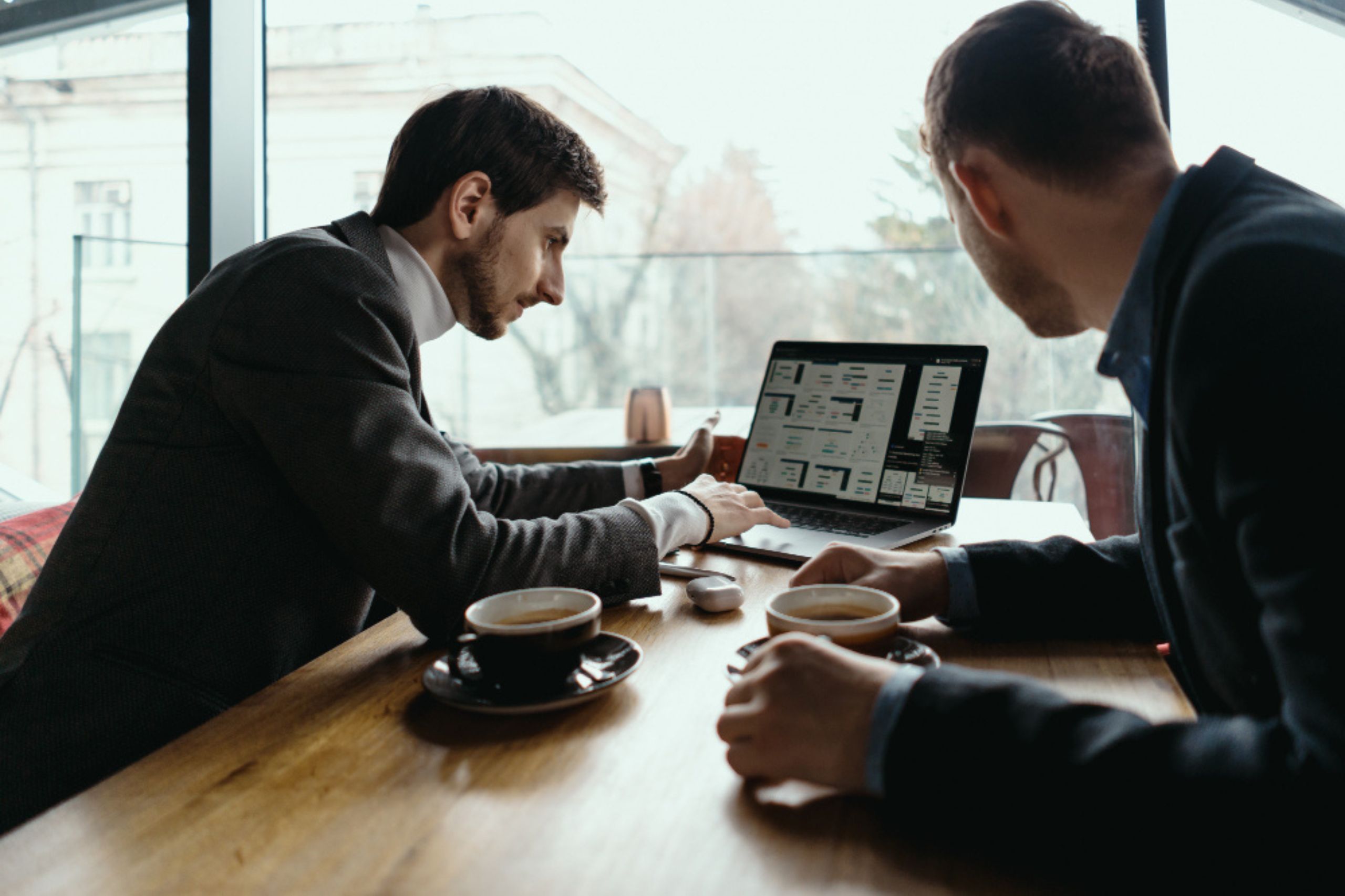 Two men in suits sit at a table with coffee, looking at a laptop displaying charts and diagrams inside a modern office or cafe.