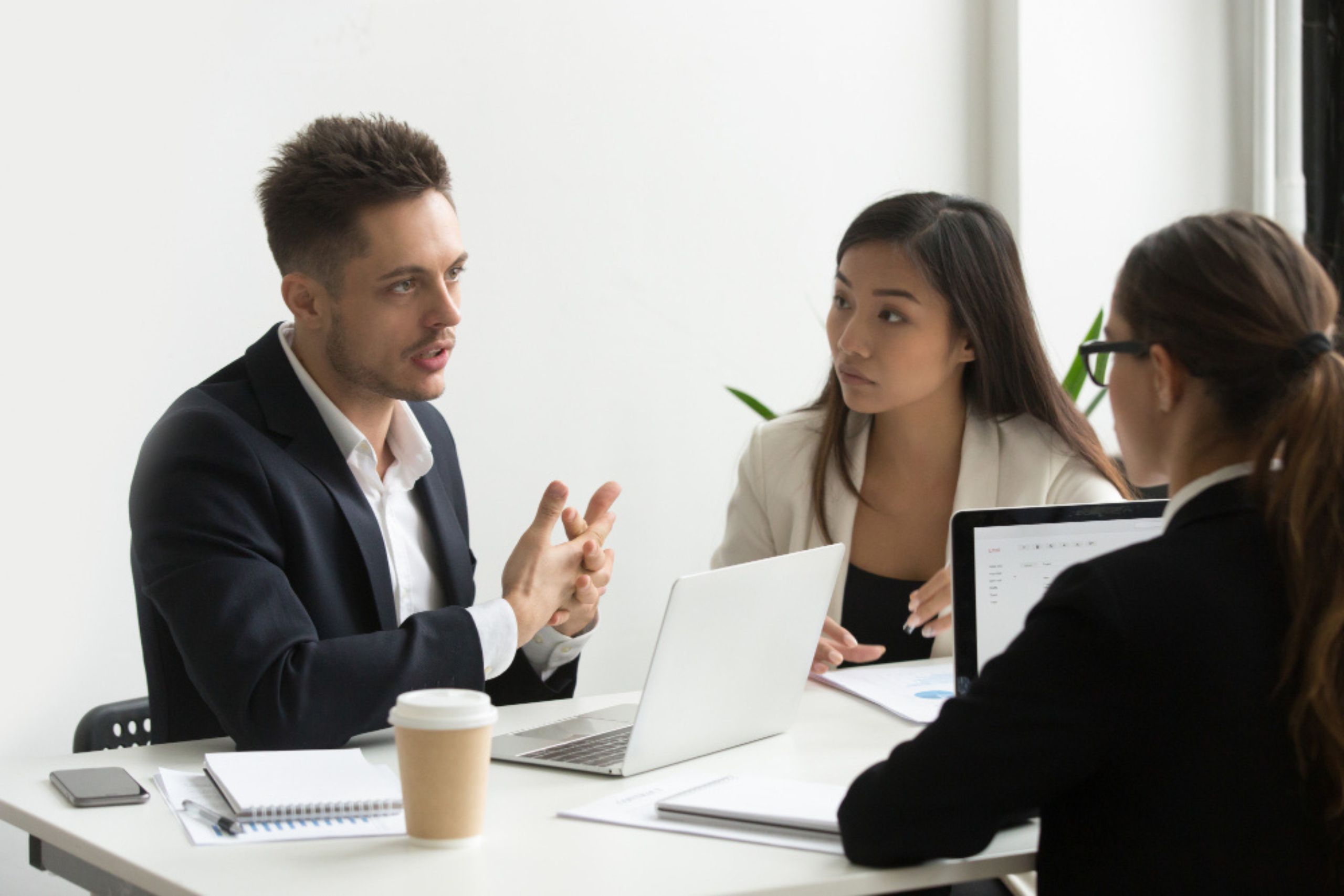 Three people in business attire sit at a table with laptops and documents, engaged in a serious discussion in an office setting.