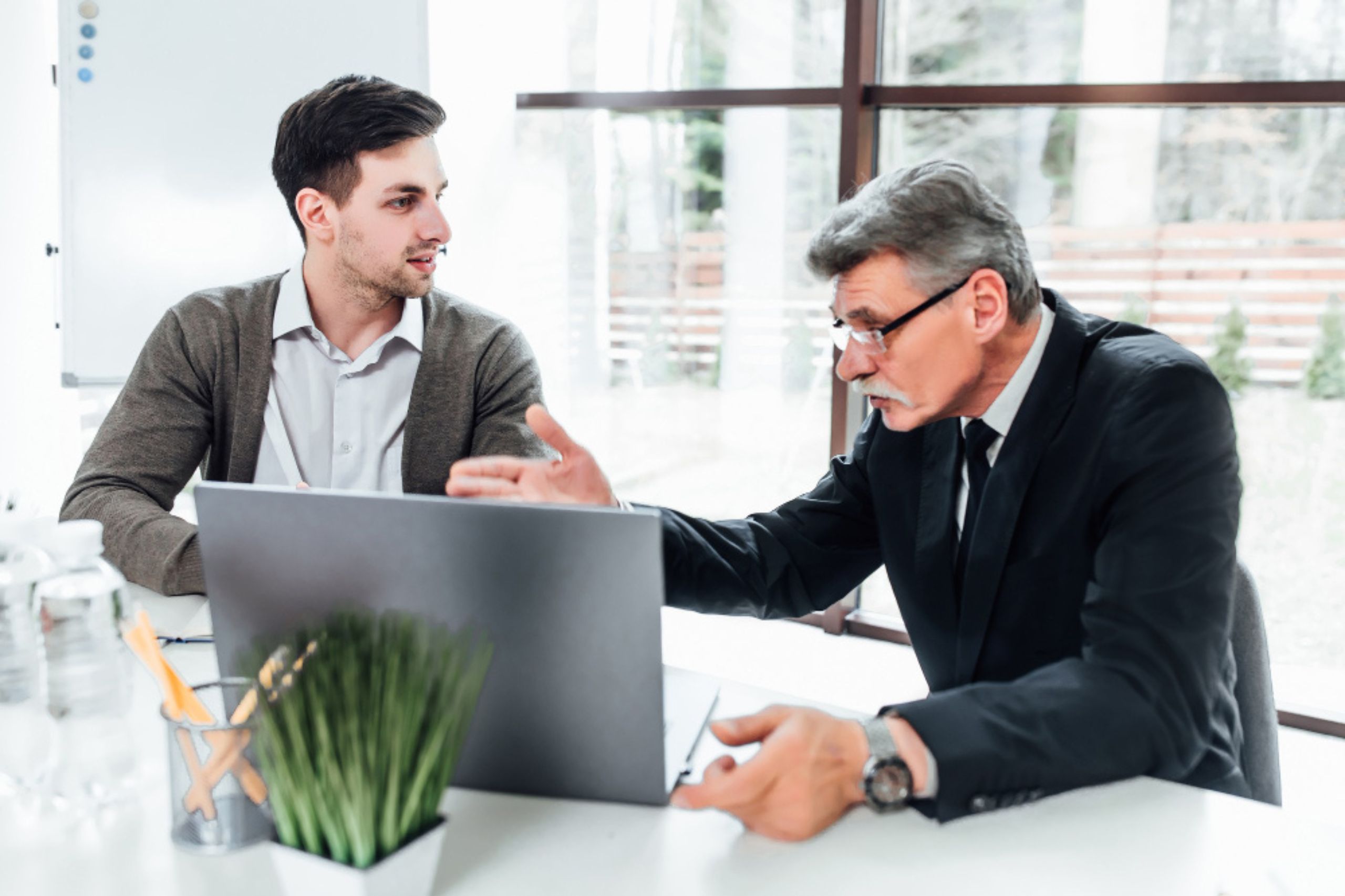 Two men sit at a desk with a laptop; one gestures while speaking, and the other listens attentively in a modern office setting.
