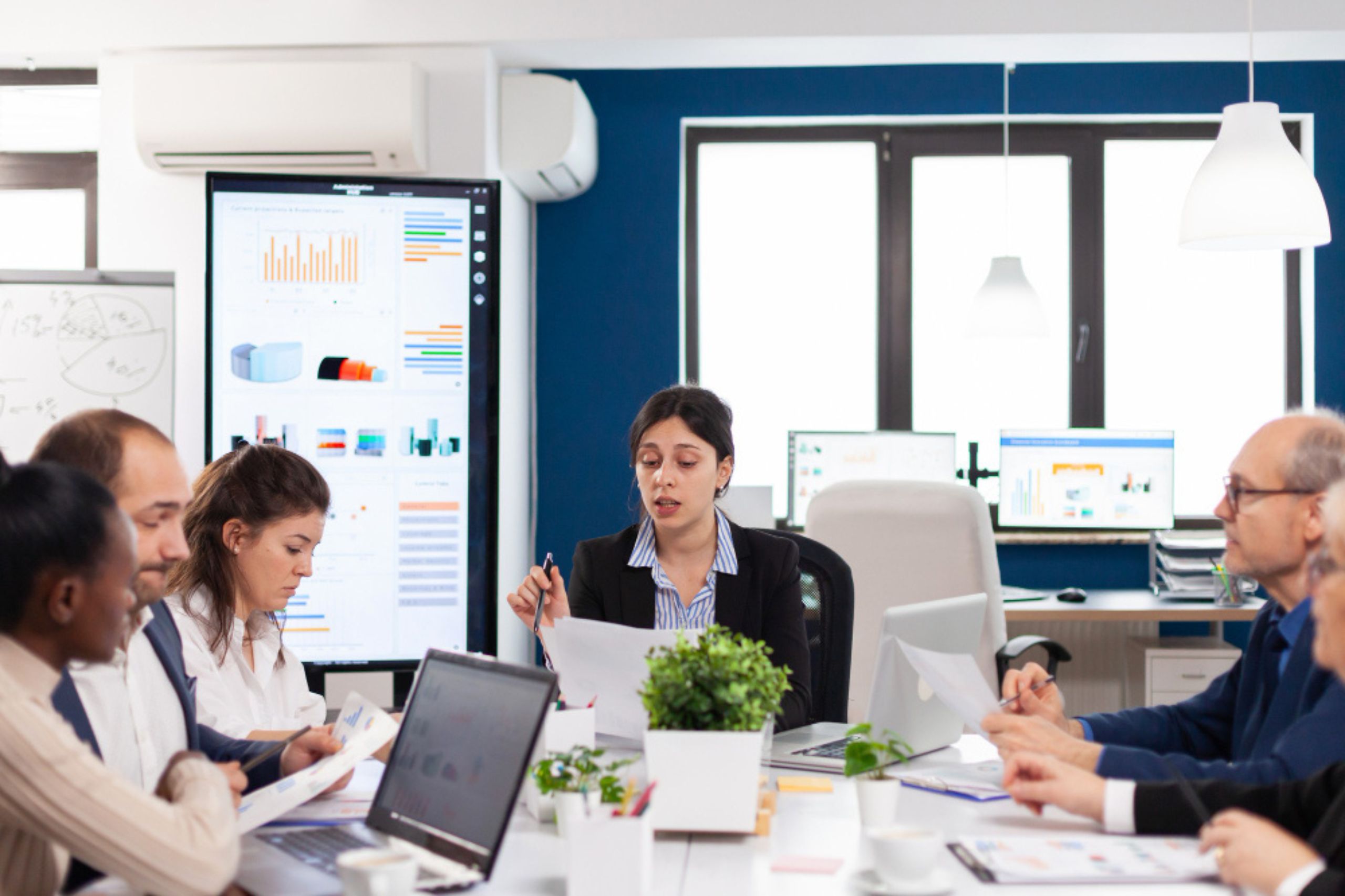 A group of professionals sits around a meeting table with laptops and papers, while a woman in the center speaks; charts are displayed on screens in the background.