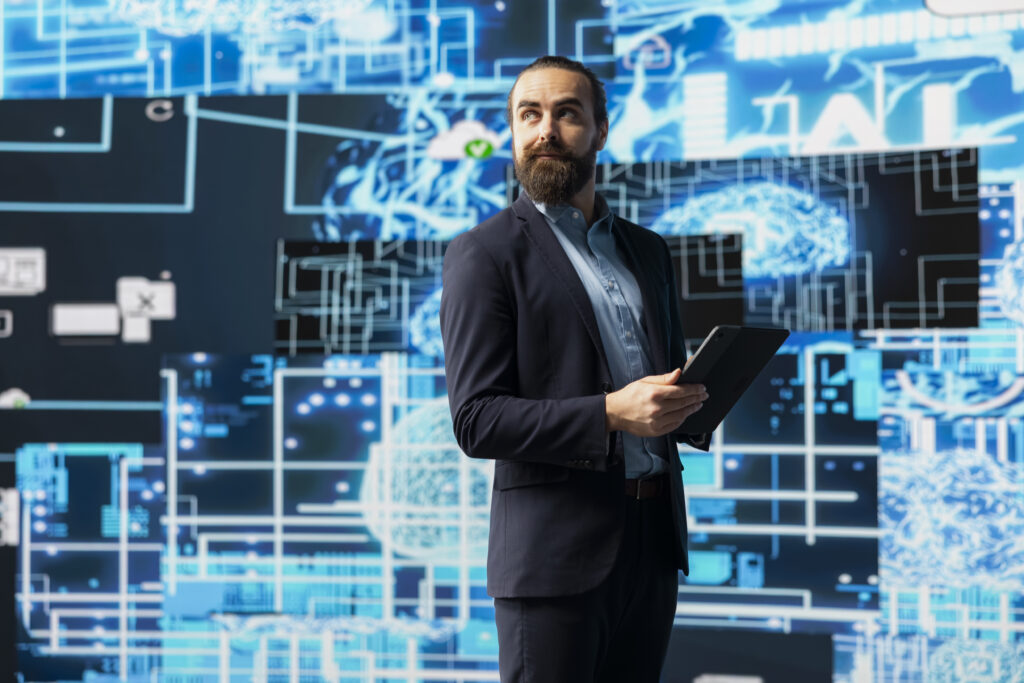 A man in a business suit holds a tablet and stands in front of a digital screen displaying abstract technology and data graphics.