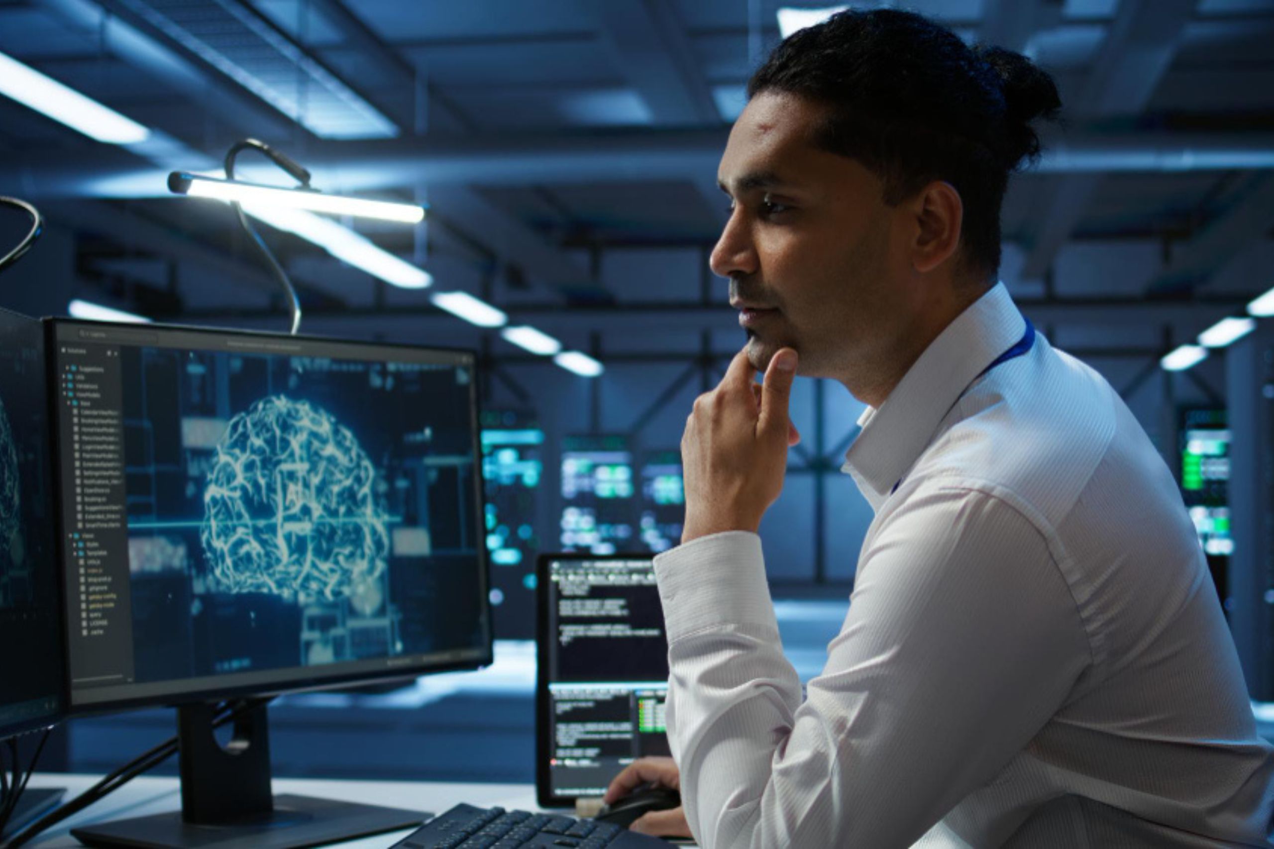 A man in a white shirt looks thoughtfully at a computer monitor displaying a digital image of a brain in a modern, high-tech office.