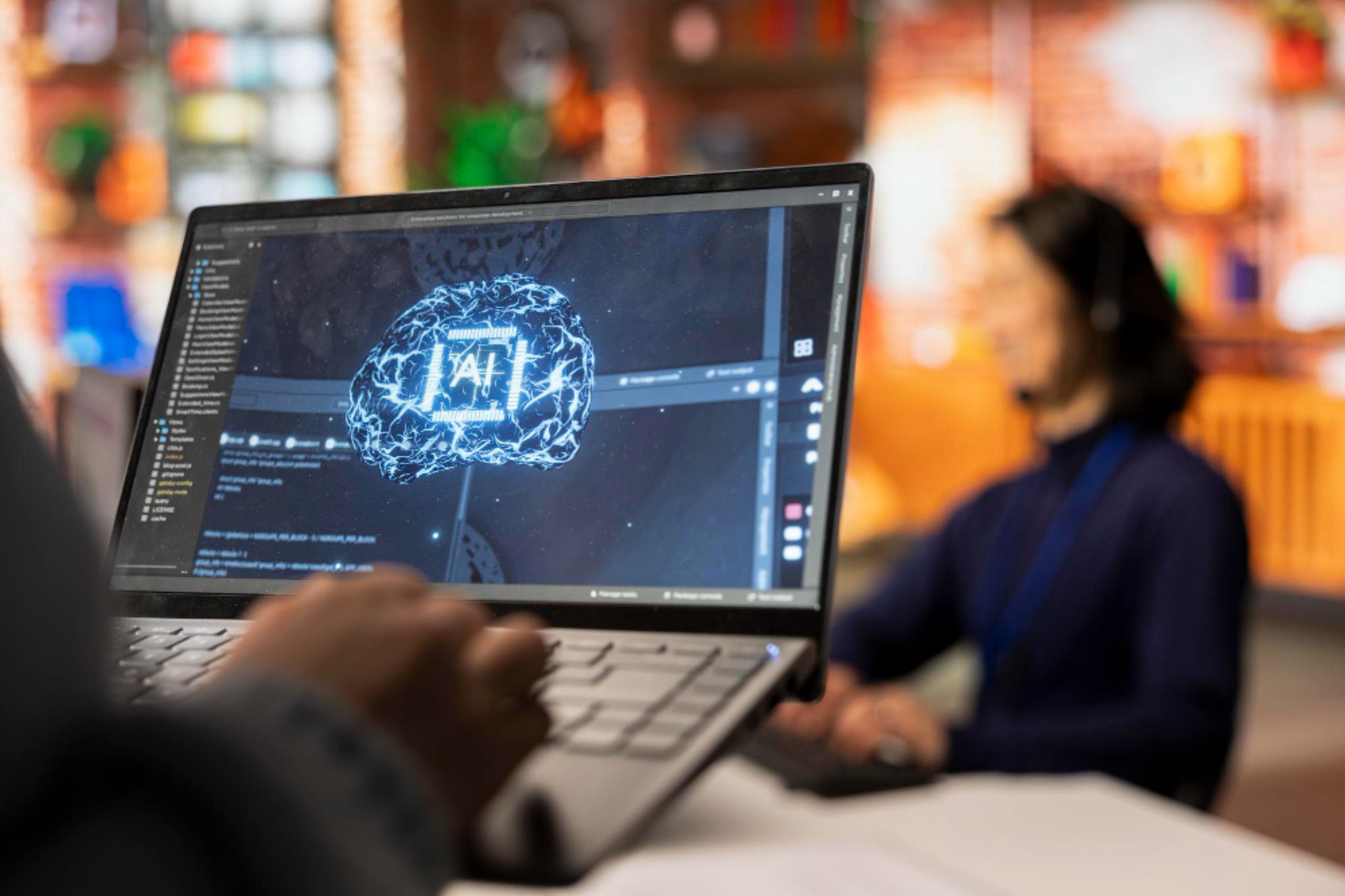 A person holds a laptop displaying a digital AI brain graphic, while another individual works in the blurred background.