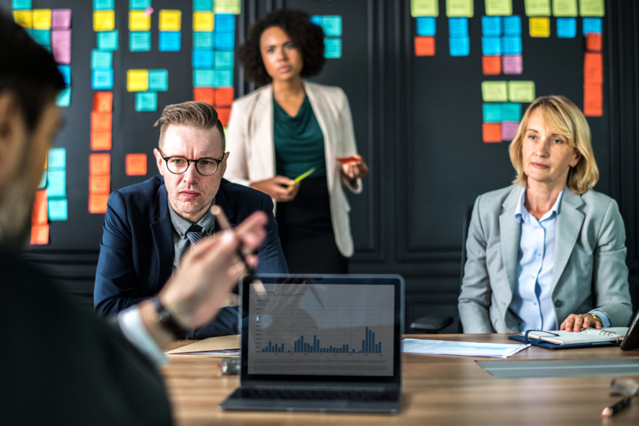 Three people in business attire sit around a table in a meeting room with colorful sticky notes on the wall and a laptop displaying a bar chart in the foreground.