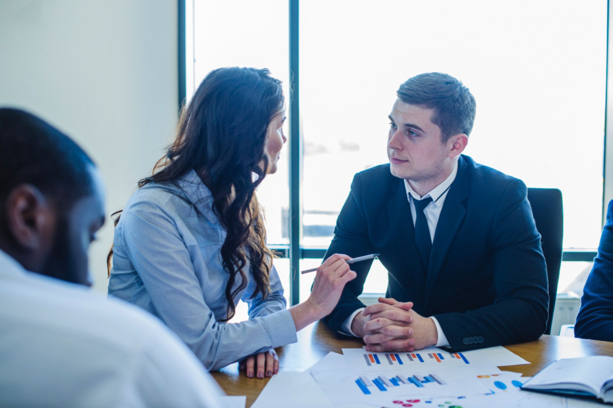 Two people in business attire sit at a table discussing documents with charts, while another person sits nearby, partially visible.