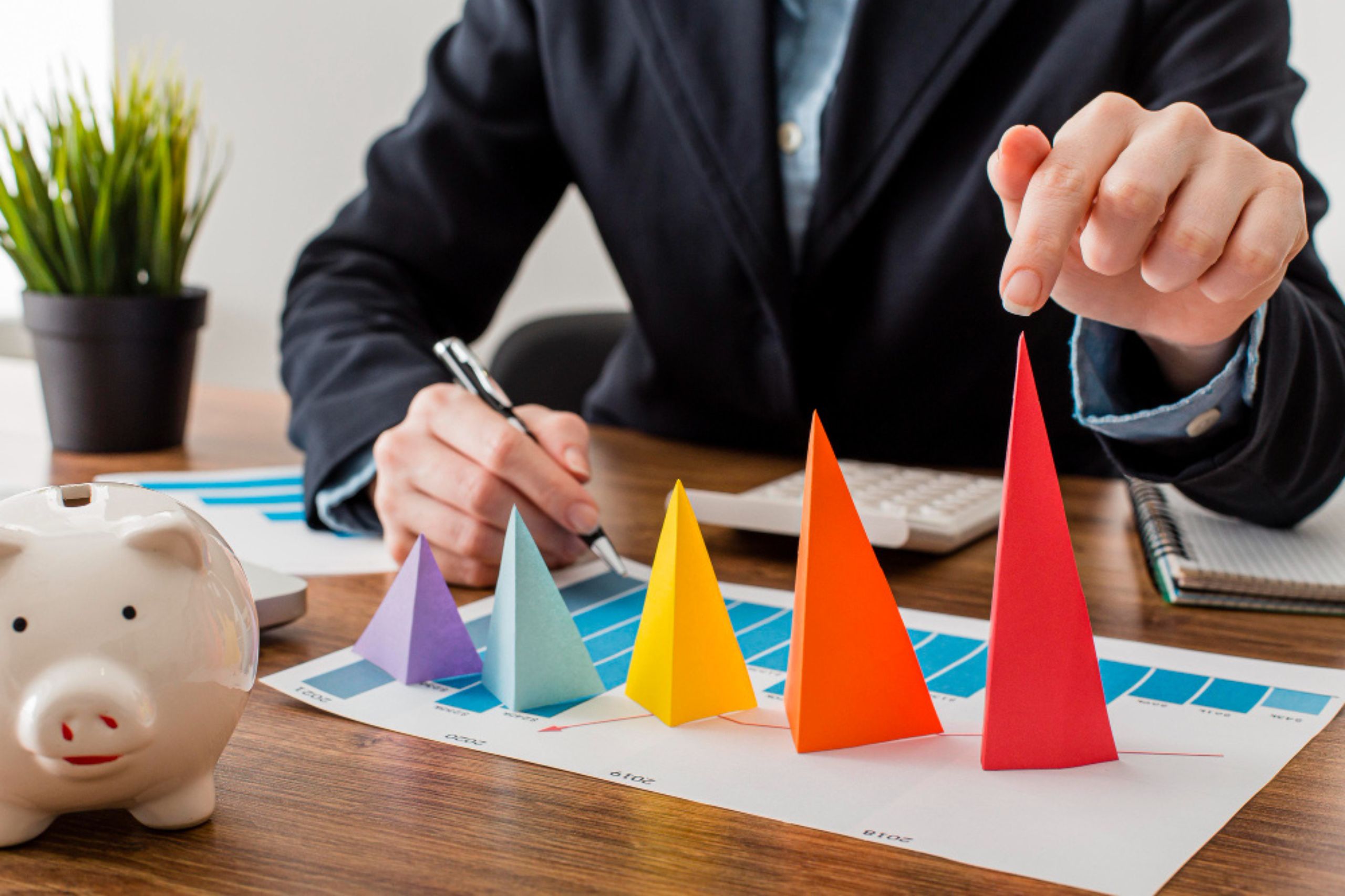 A person in business attire analyzes colorful 3D pyramid charts on a desk with a piggy bank, documents, notebook, and calculator.