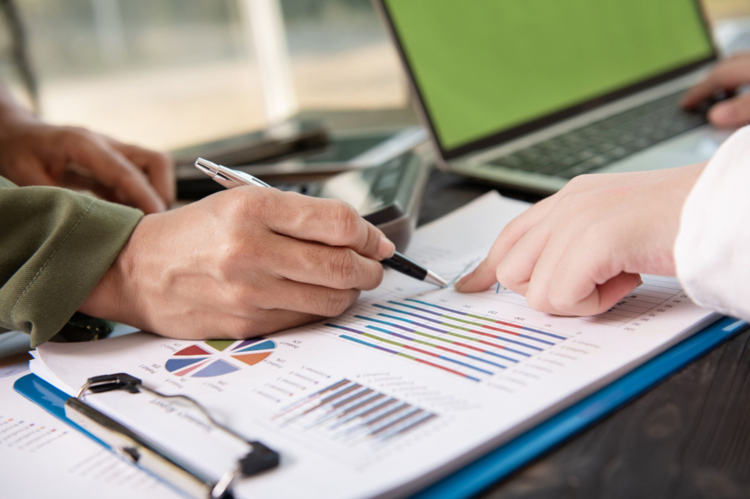 Two people review printed charts with graphs and pie charts at a desk, using pens to point at data, with a laptop and clipboard nearby.