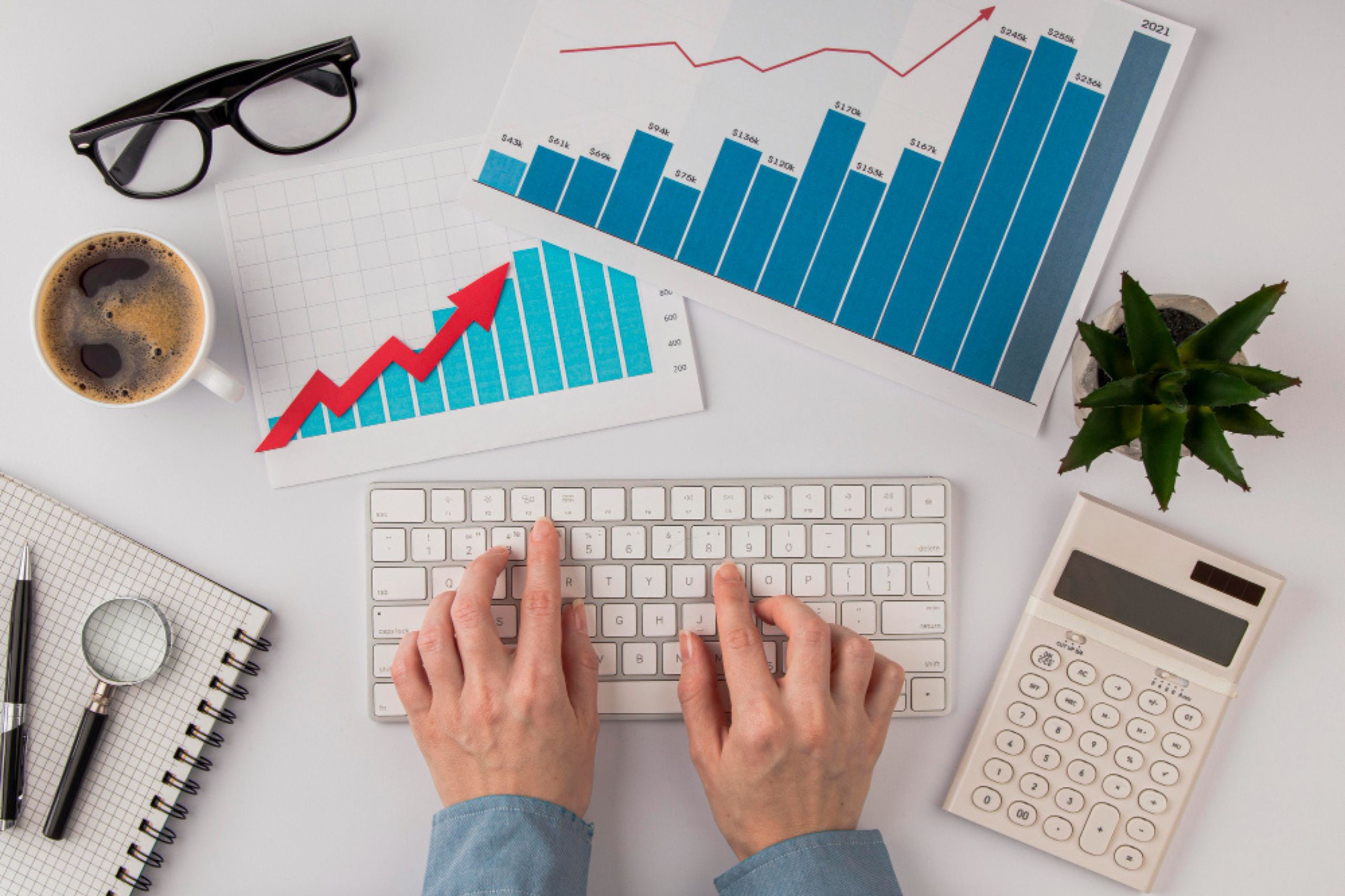 Hands typing on a keyboard with bar and line graphs, calculator, notepad, coffee, glasses, and a small plant on a white desk.