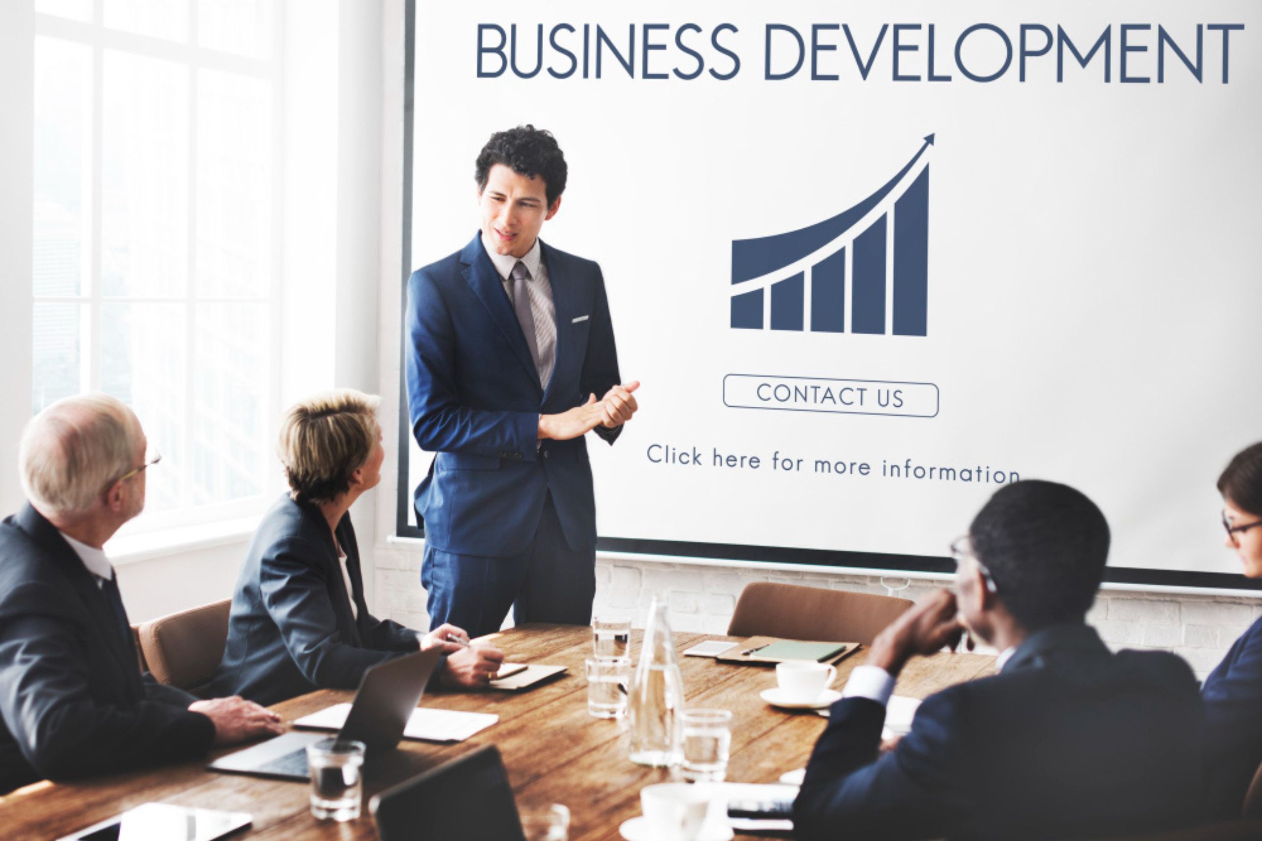 A man in a suit presents to four colleagues in a conference room with a screen displaying "Business Development" and a growth chart.
