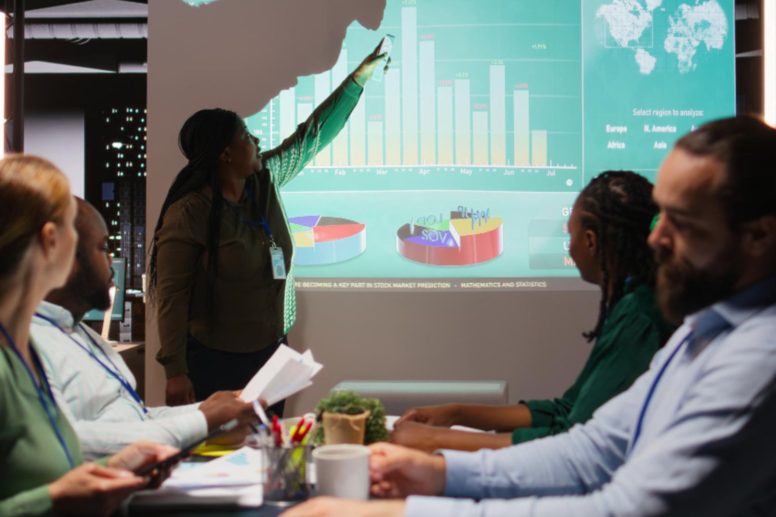 A person gives a presentation with graphs and charts projected on a screen while four colleagues sit at a table and listen attentively.