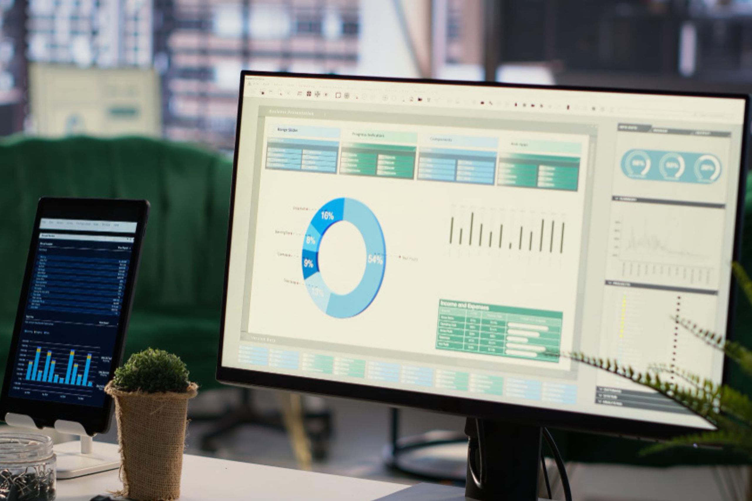 A computer monitor displays charts and graphs on a dashboard, while a tablet shows data analytics. A small potted plant sits on the desk in the foreground.