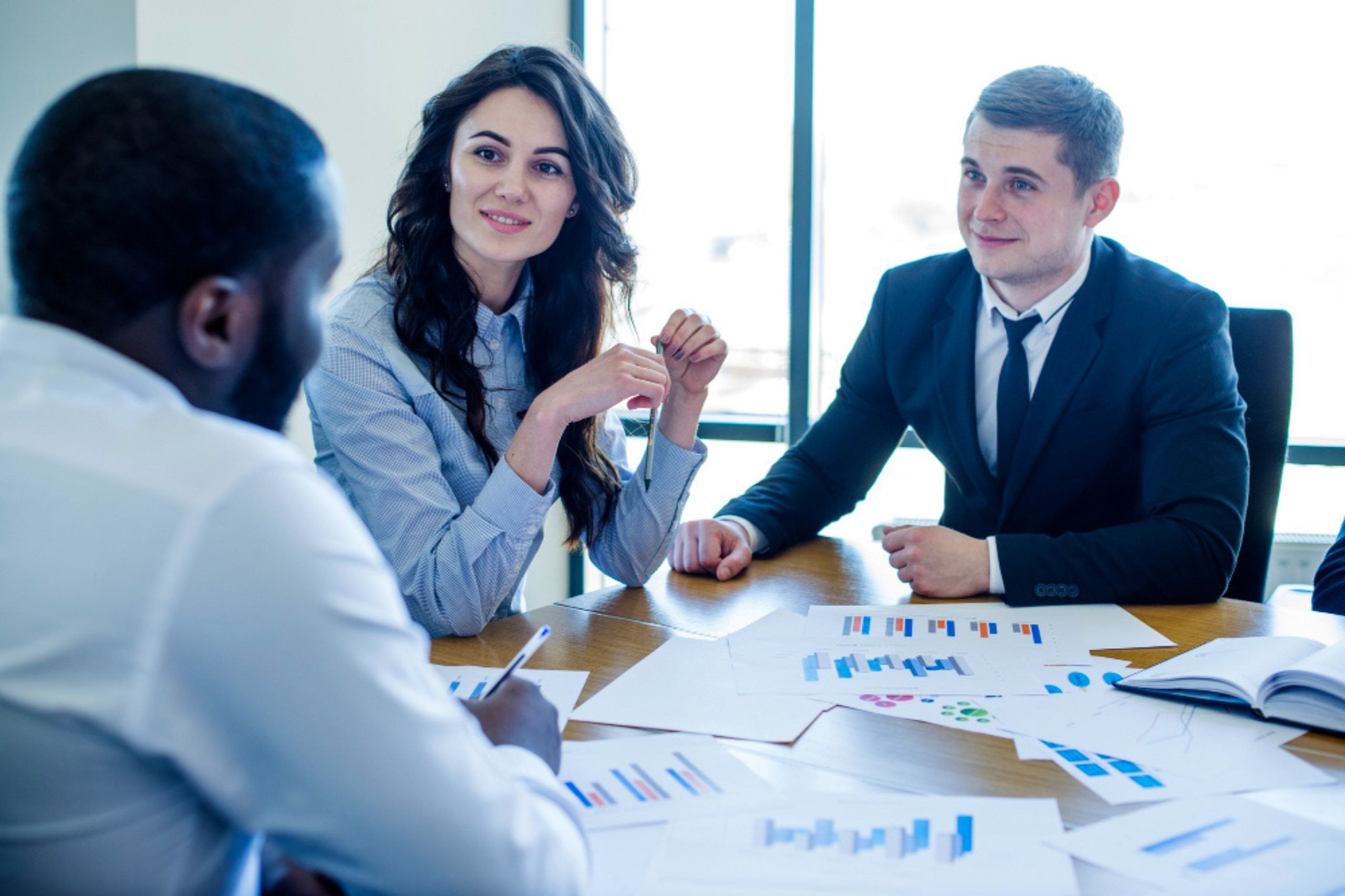 Three people in business attire sit at a table discussing documents and charts during a meeting in a well-lit office.
