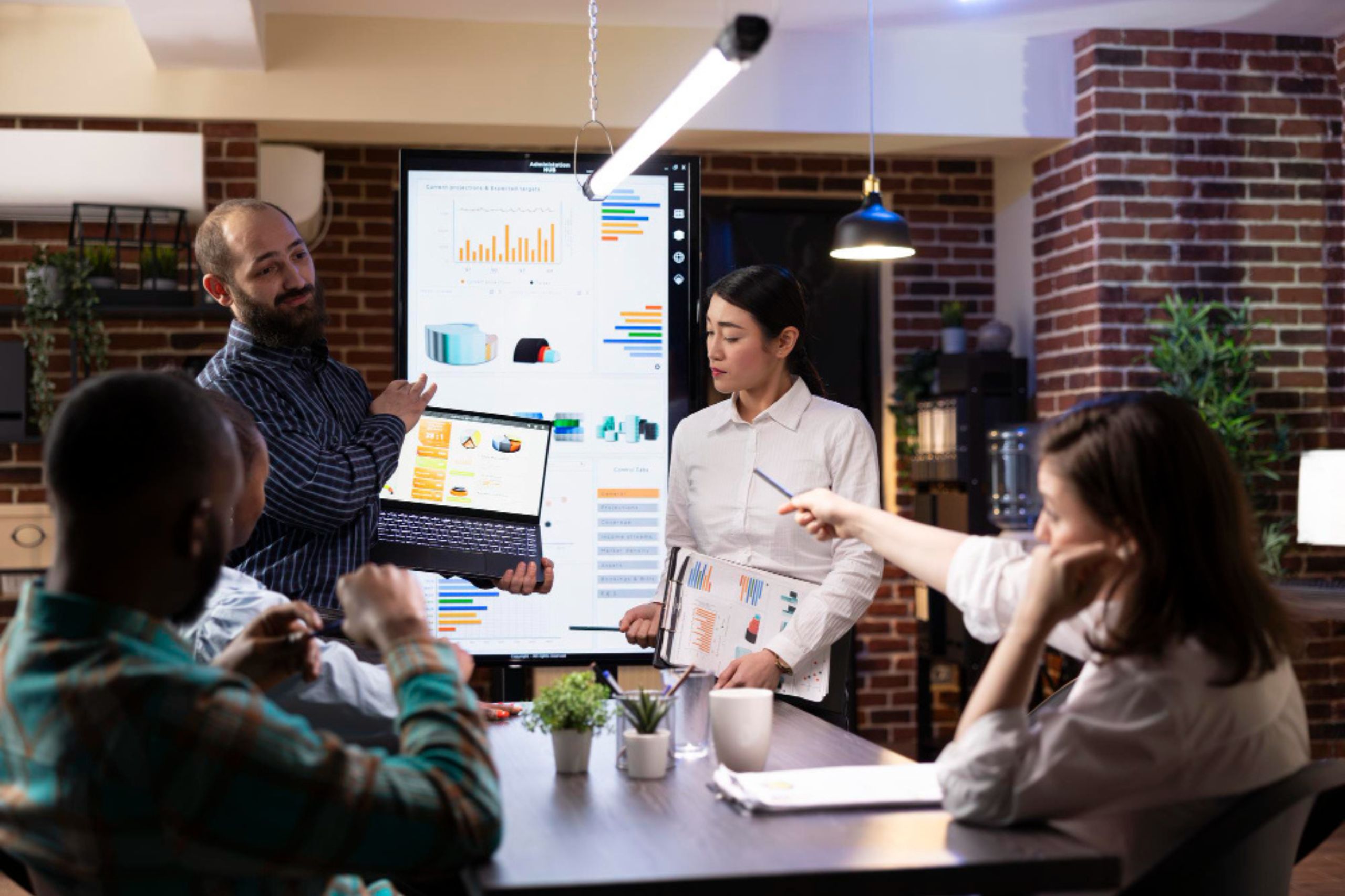 Four people in a modern office discuss data displayed on a laptop and a large screen with charts and graphs during a meeting.