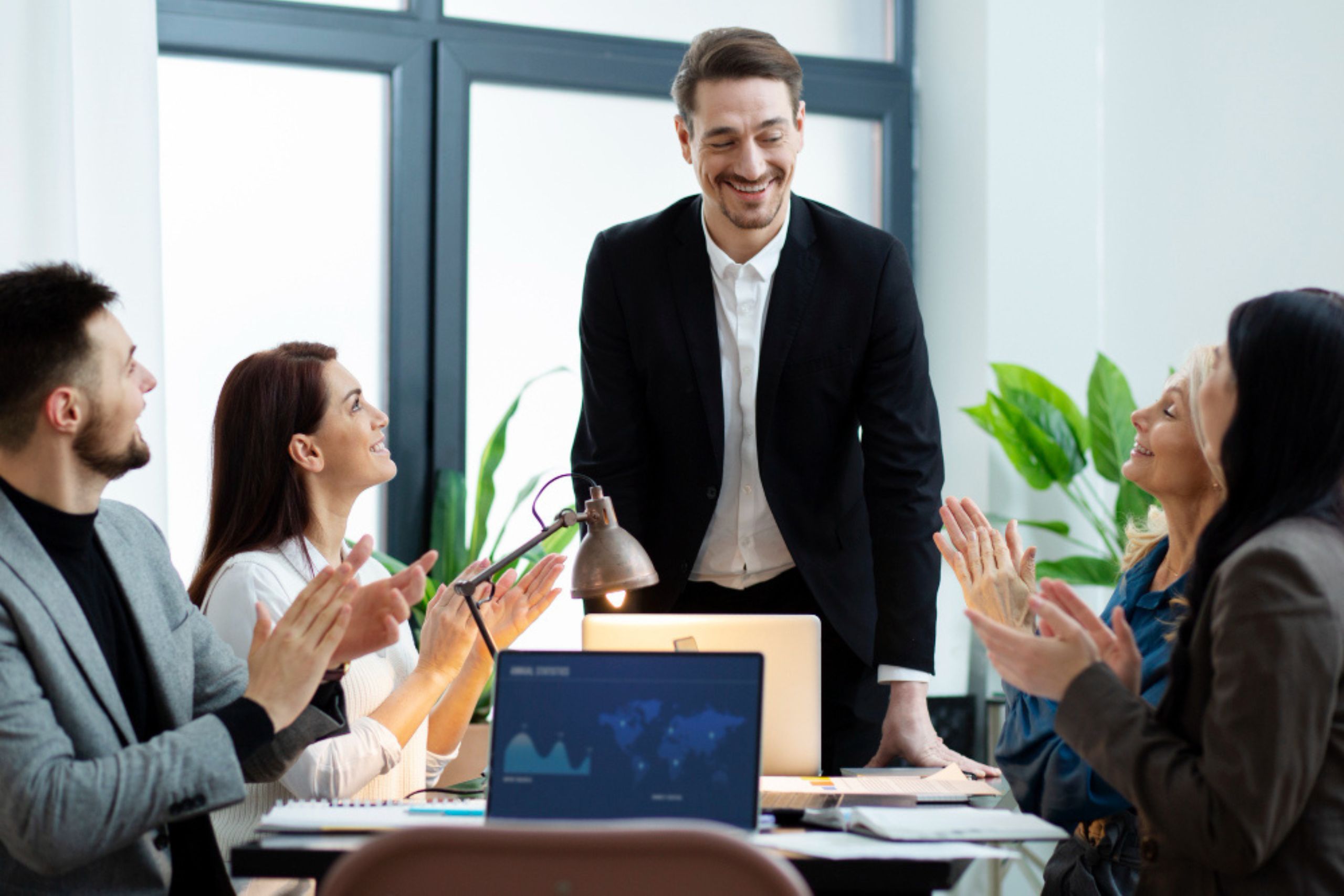 Four people sit around a table applauding a man in a suit who is standing and smiling. Laptops, plants, and paperwork are visible in the office setting.