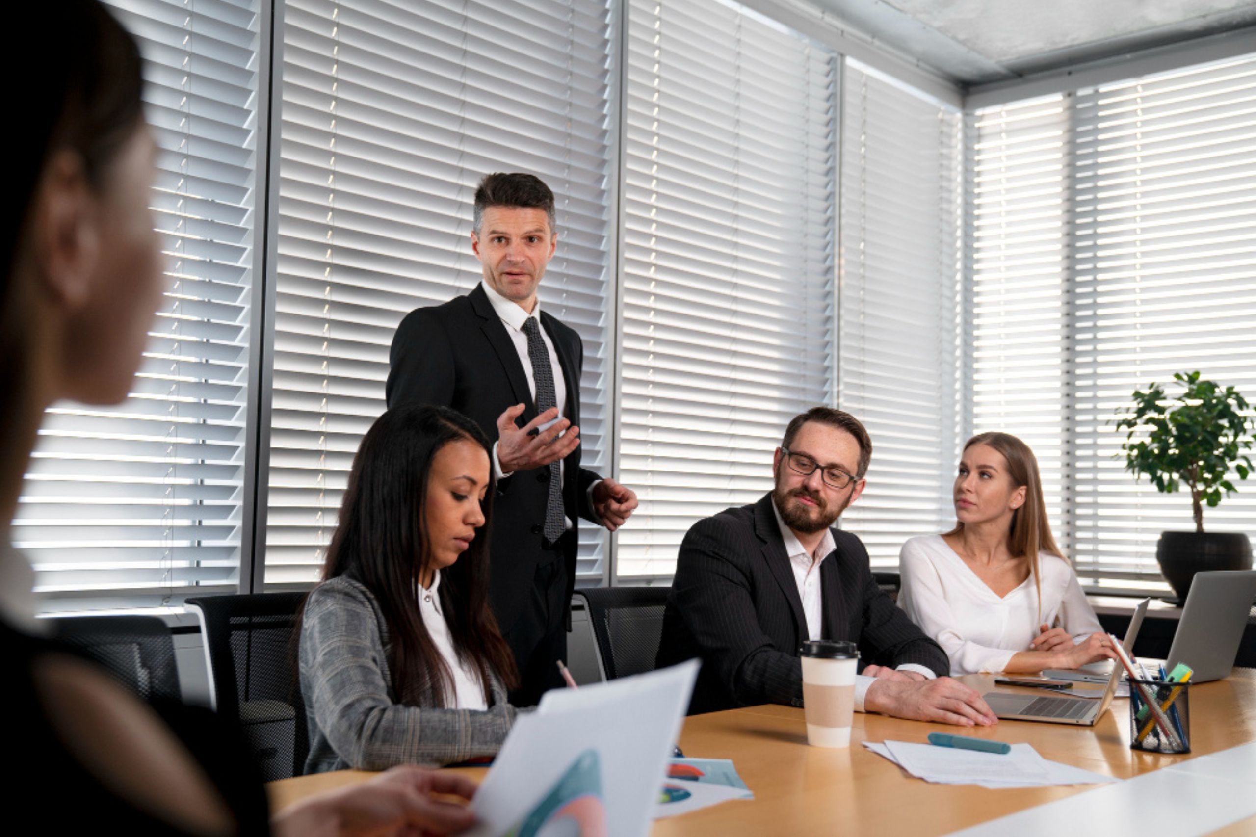 A man in a suit stands and speaks during a business meeting while three colleagues sit at the conference table, listening.