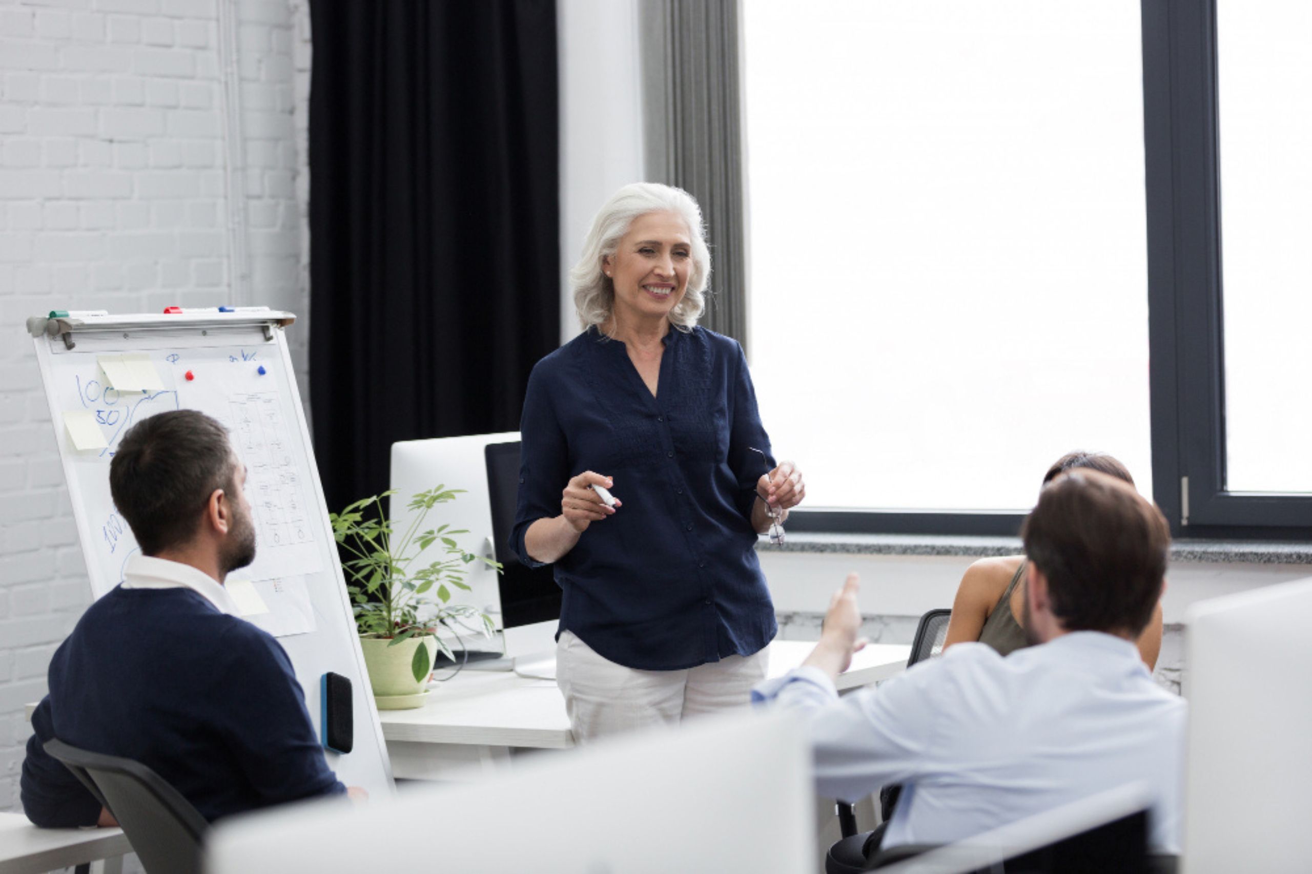 An older woman stands and presents to three seated colleagues in an office meeting, with charts and notes visible on a flip chart.