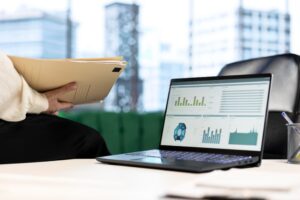 A person holding file folders sits beside a desk with an open laptop displaying various business charts and analytics.