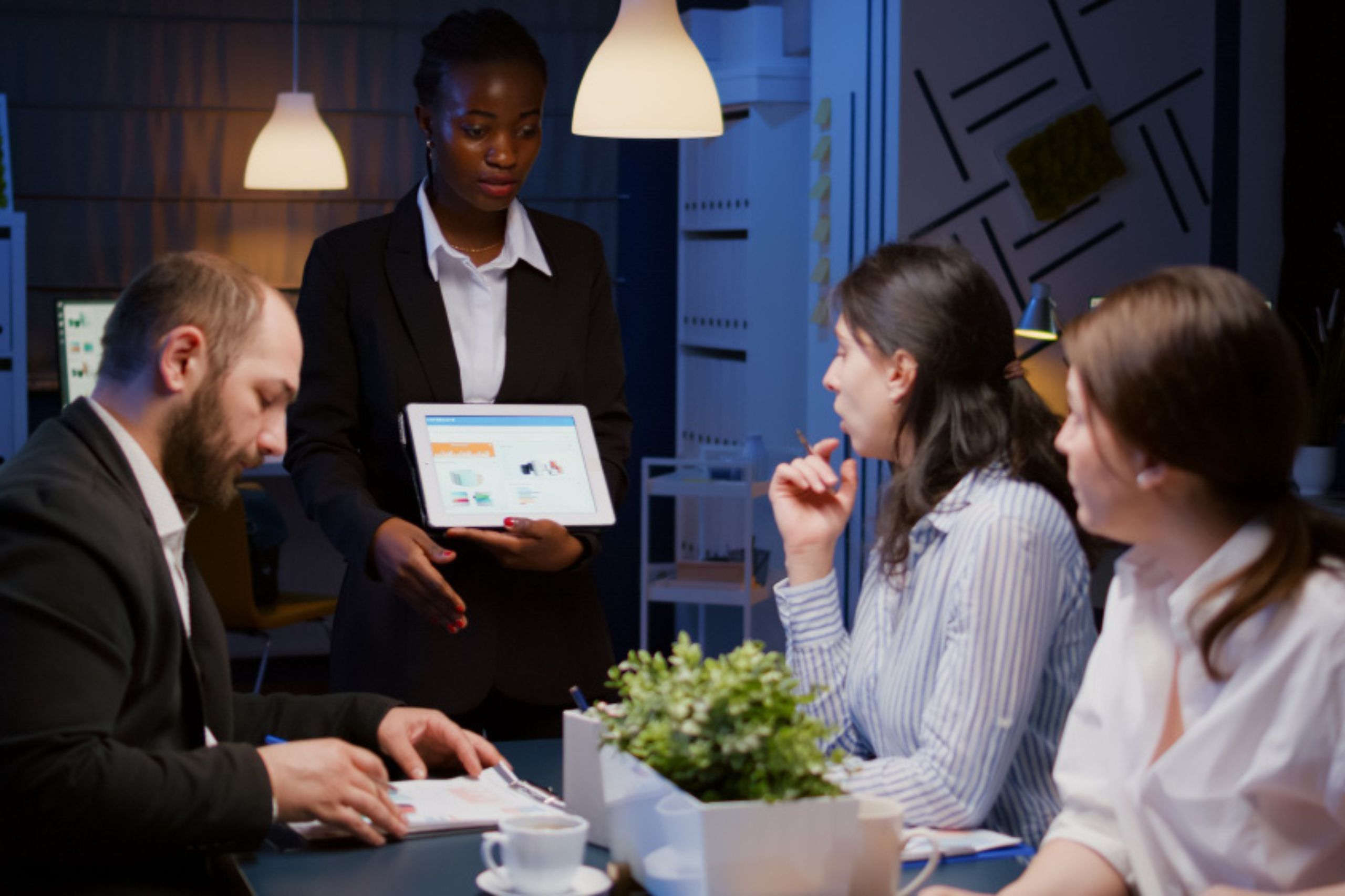 A woman standing and holding a tablet presents data to three colleagues seated at a table in an office meeting room.