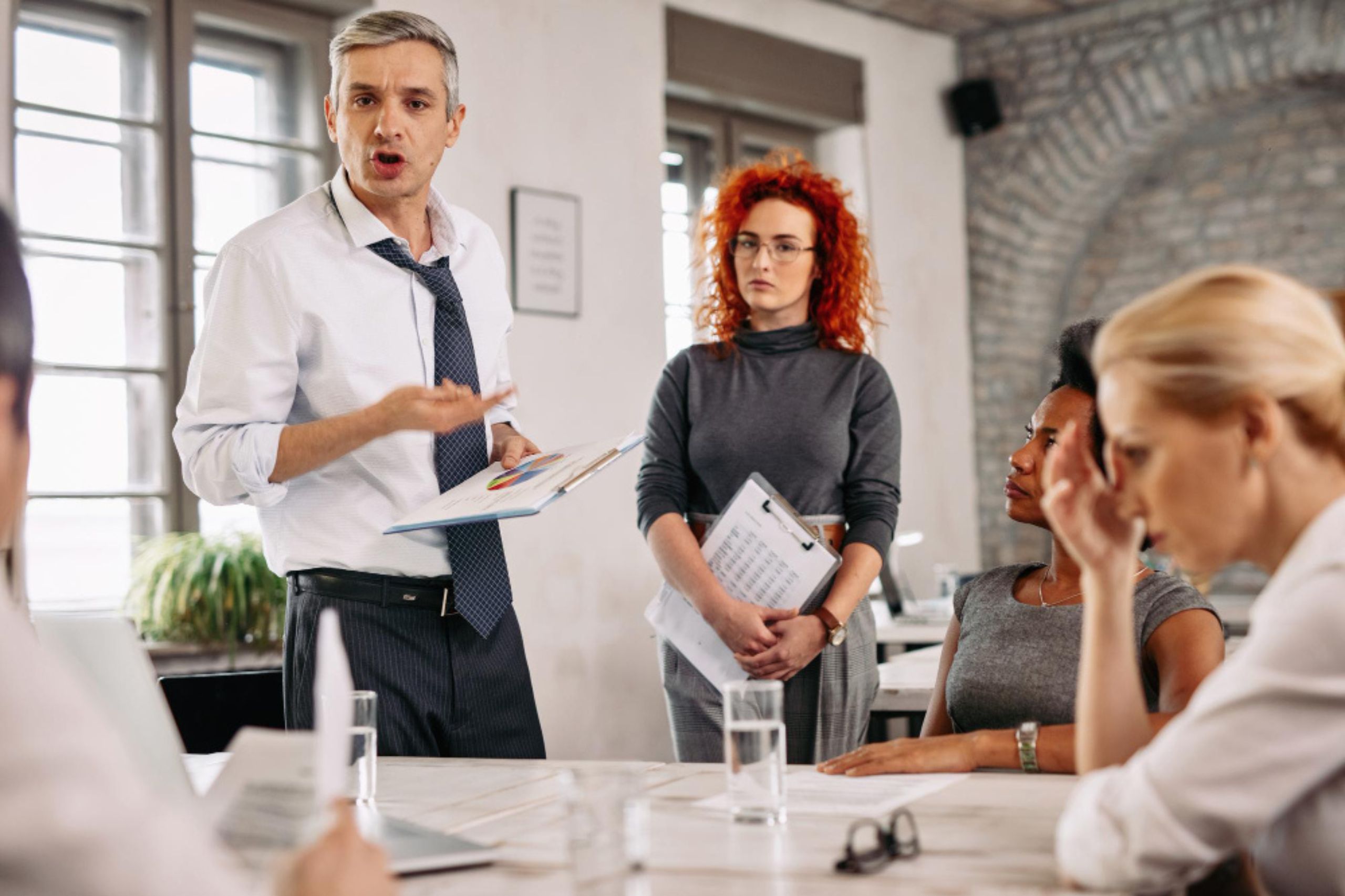 A man in a white shirt speaks emphatically to colleagues during a meeting, while others listen; documents and glasses of water are on the table.