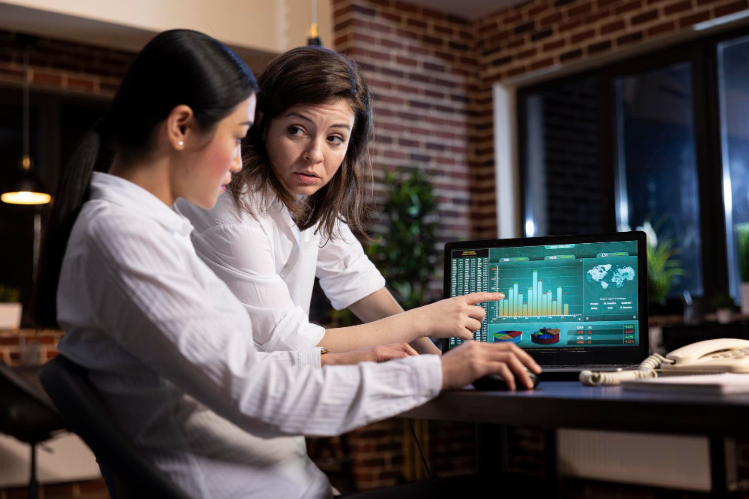 Two women at a desk review data on a laptop screen displaying charts and graphs in an office setting.