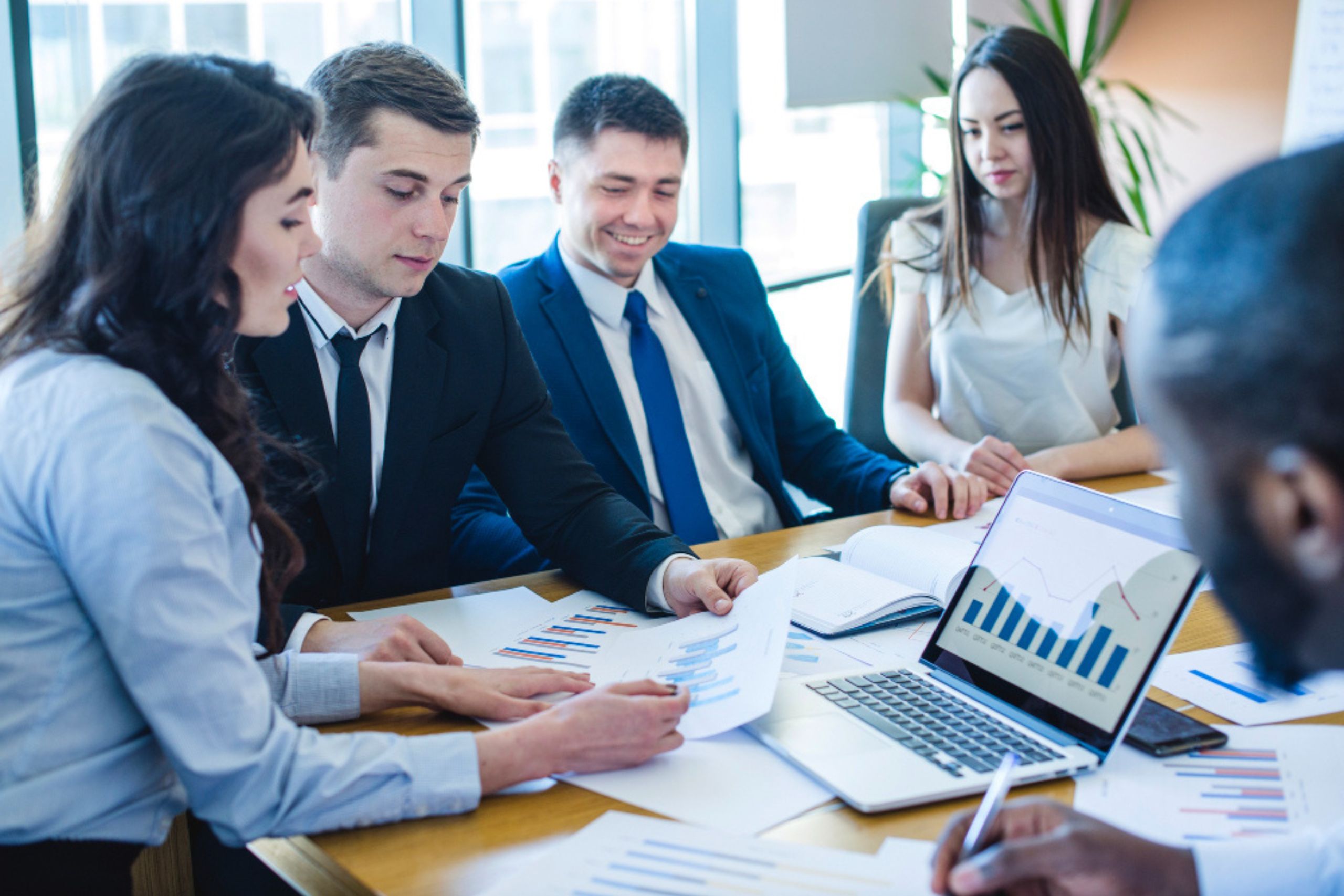 Four business professionals in formal attire review charts and graphs at a meeting table, with a laptop displaying a bar graph in front of them.
