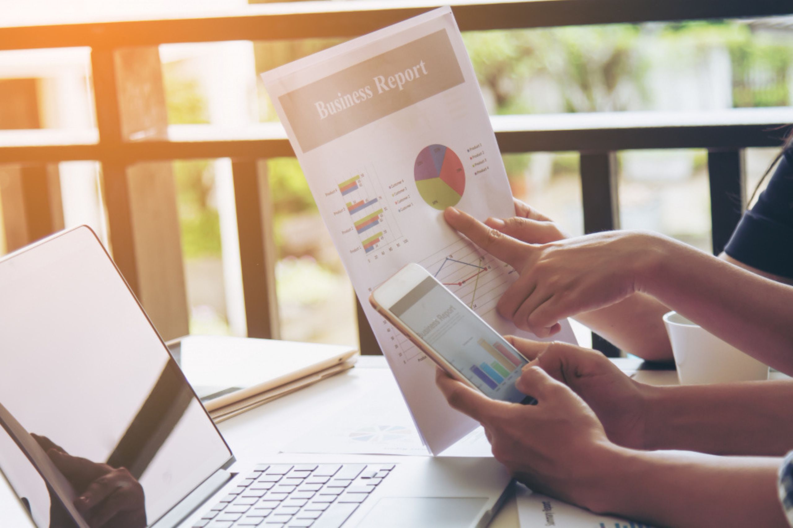 Two people review a printed business report with charts and graphs at a table with a laptop and smartphone.